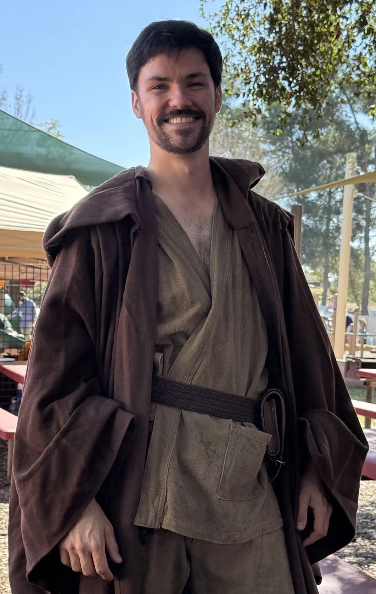 A man dressed as a Jedi from Star Wars, wearing a brown robe and beige tunic, standing outdoors with trees and tents in the background, smiling at the camera.