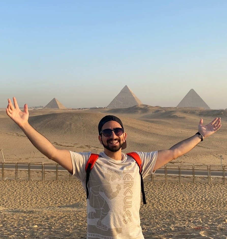 A man with sunglasses, a black cap, and a red backpack smiling with his arms raised in front of the pyramids in Egypt.