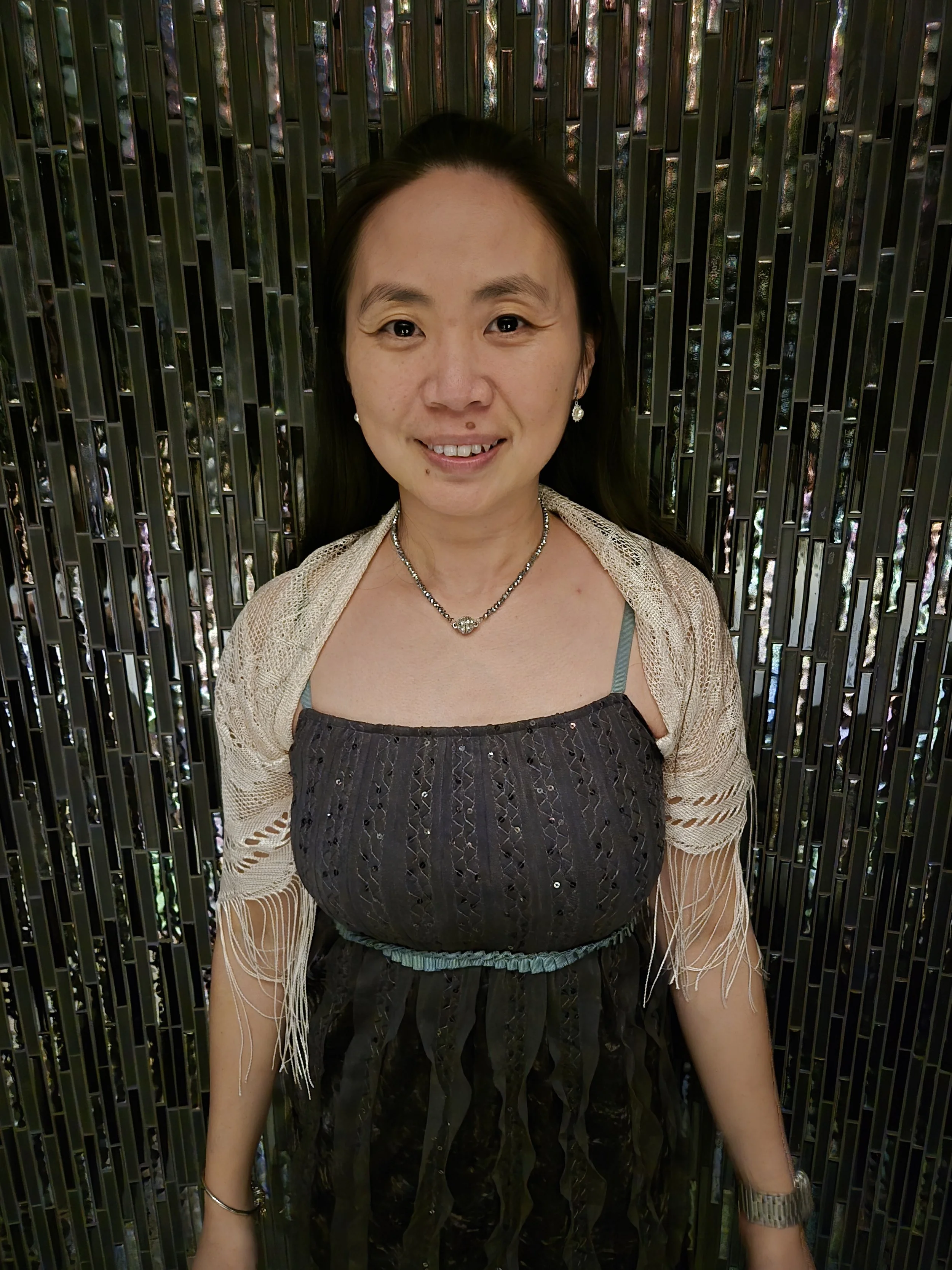 A woman with long dark hair smiling at the camera, standing in front of a reflective, patterned metallic background.