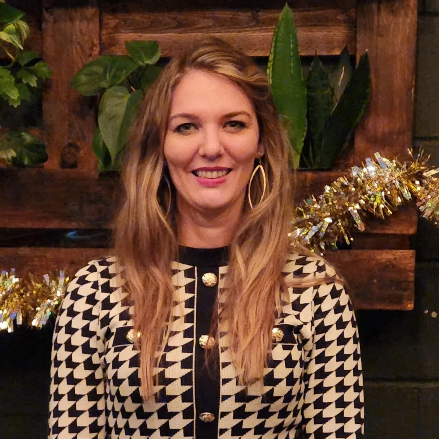 A woman with long wavy blonde hair wearing large gold hoop earrings, a black and beige houndstooth patterned top with gold buttons, smiling in front of a wooden wall with green plants and gold tinsel decorations.