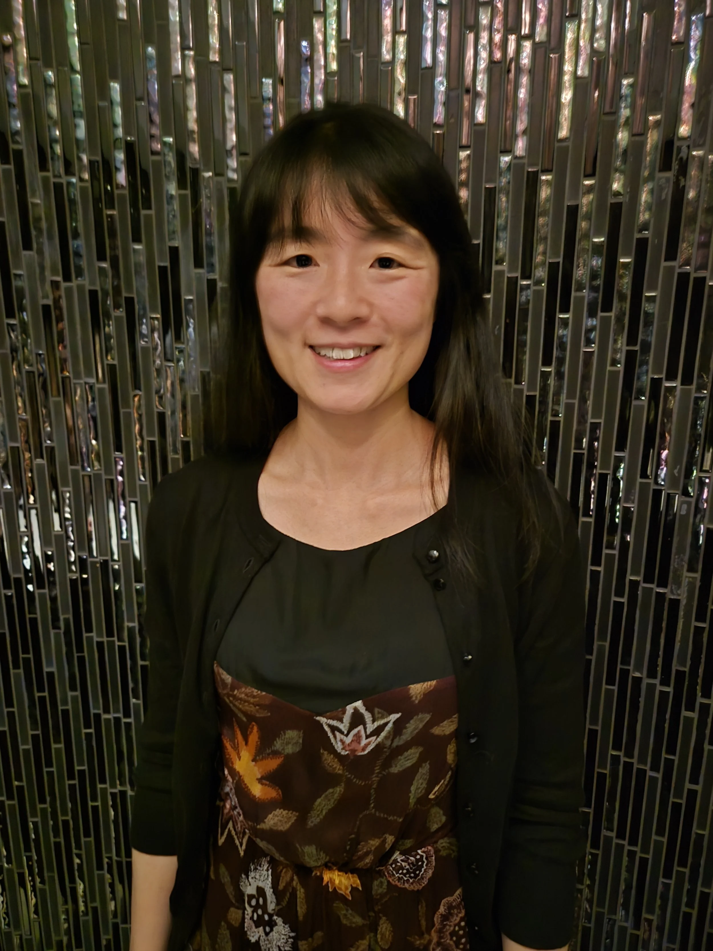 A woman with long black hair smiling in front of a shiny, metallic, patterned wall.