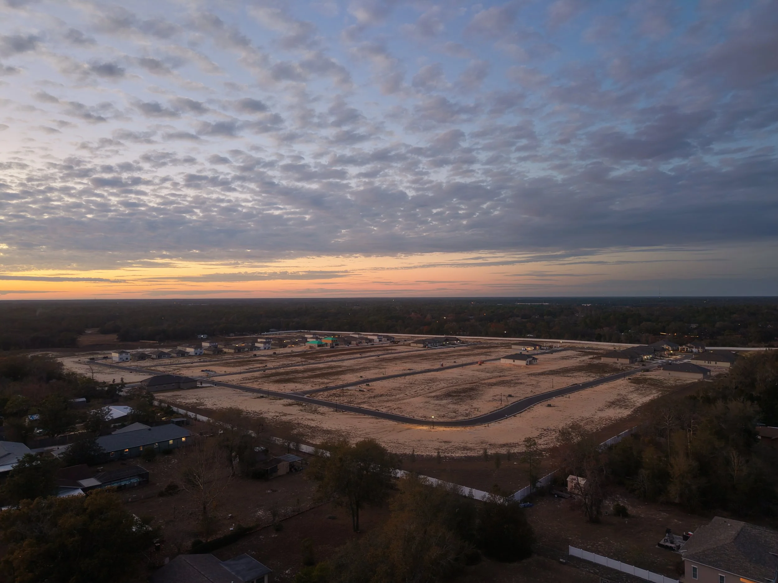 A wide view of a residential neighborhood with houses and trees in the foreground. In the distance, an empty lot with paved roads and buildings under construction. The sky is partly cloudy with the sun setting behind the horizon.