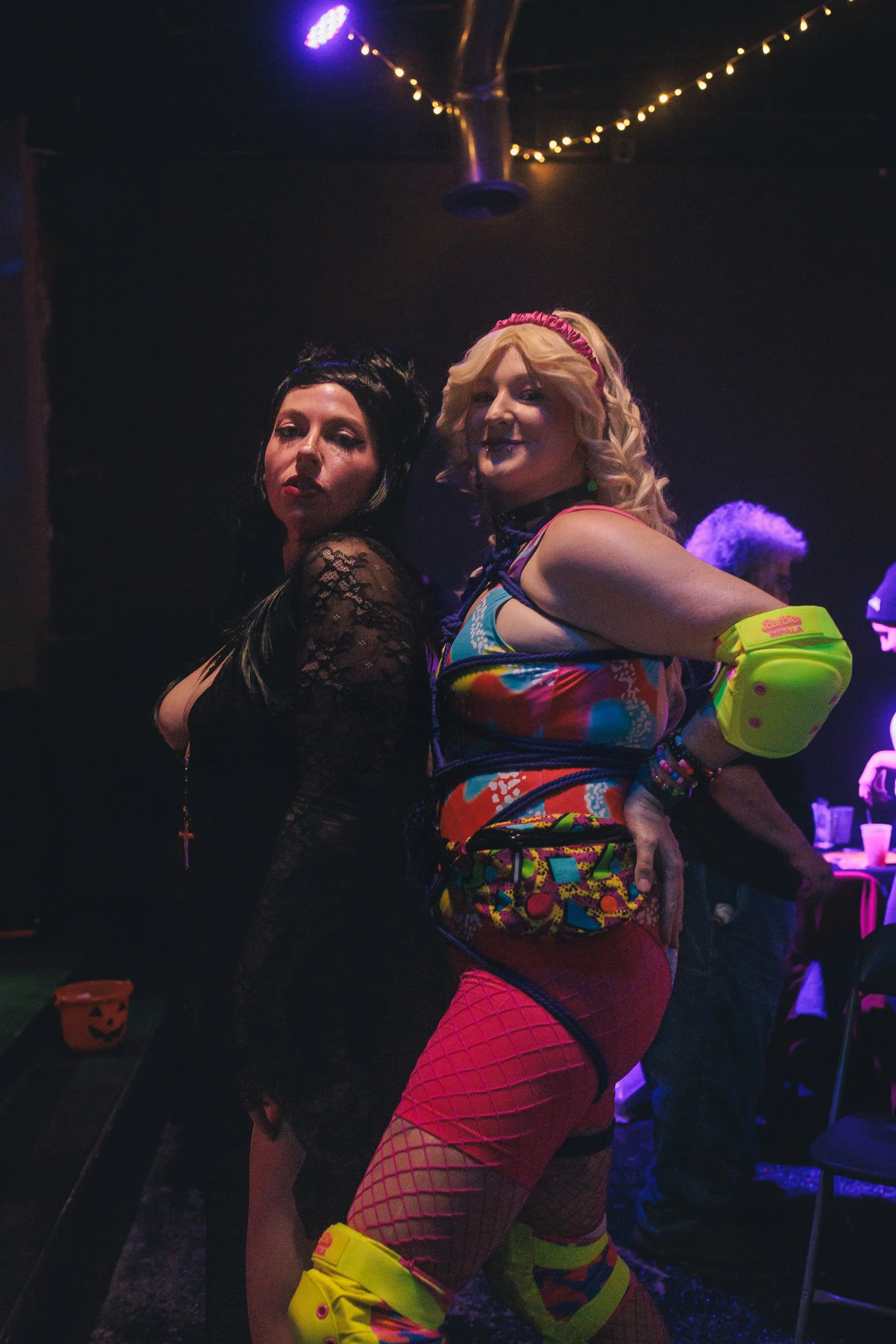 Two women in colorful costumes posing at a party with Halloween decorations, including a pumpkin bucket, in a dimly lit room with purple lighting and string lights overhead.