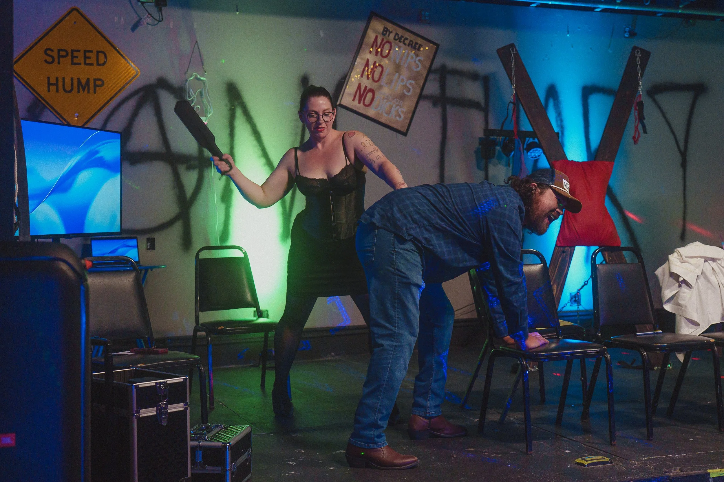 Two people are performing a game or activity on a stage with chairs, equipment, and banners in the background, in a dimly lit room with colorful lighting.