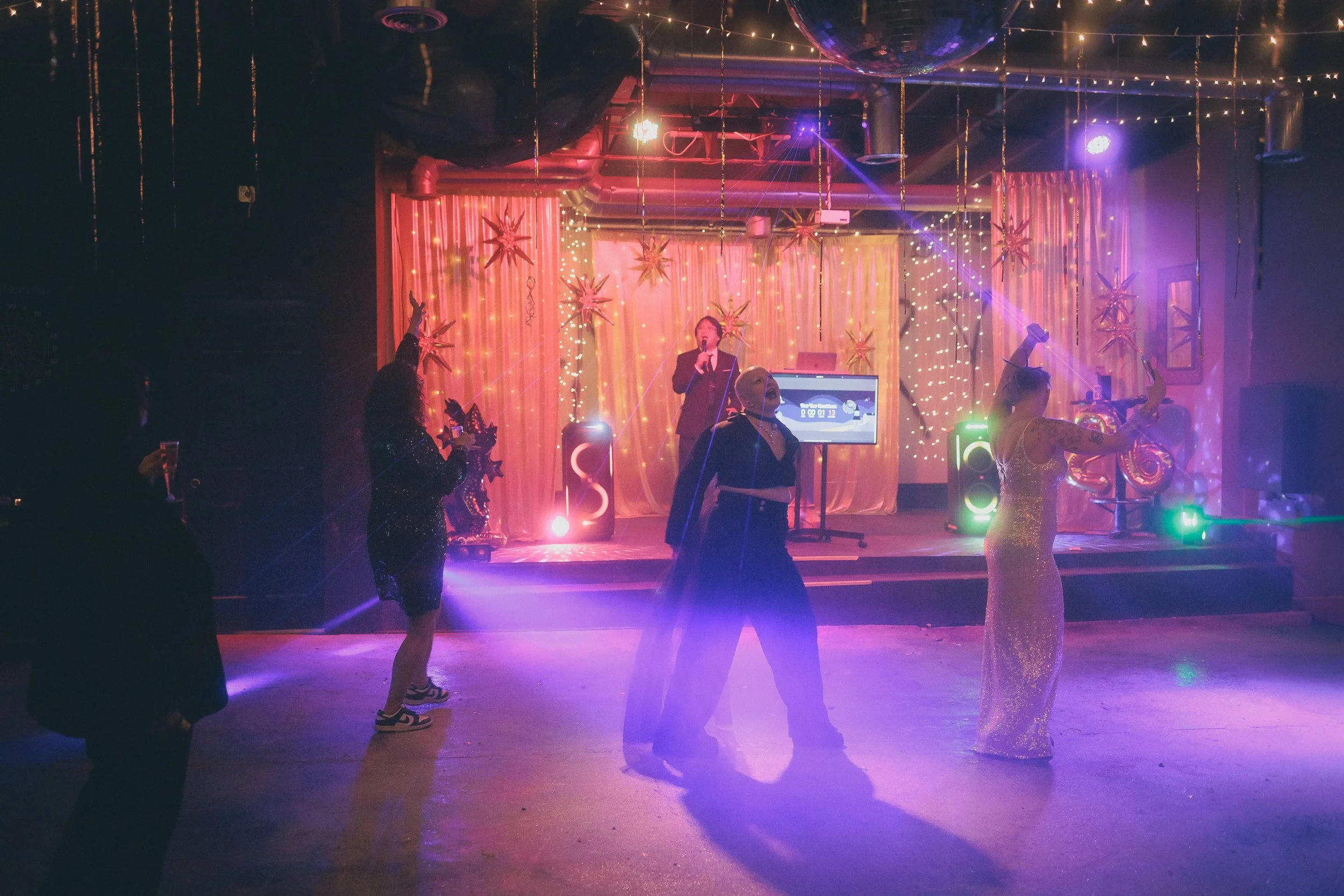 People dancing on a stage with DJ and singer, decorated with star-shaped ornaments, string lights, and a colorful light display at a party or celebration.