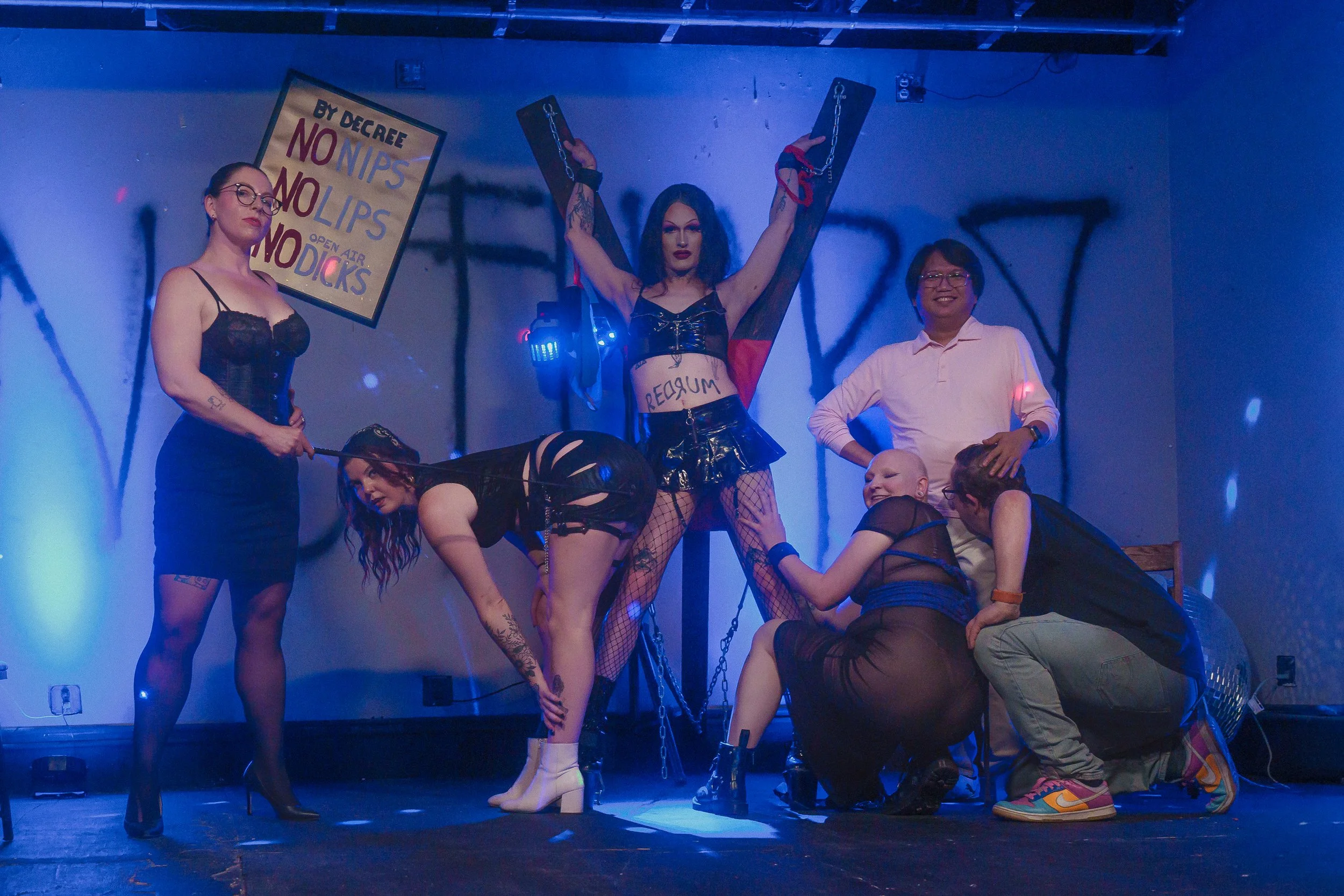 A group of women in provocative outfits on a stage, with one woman in the center wearing a black crop top and shorts with fishnet stockings, holding chains, and surrounded by others posing suggestively; some women are kneeling and others standing, wi