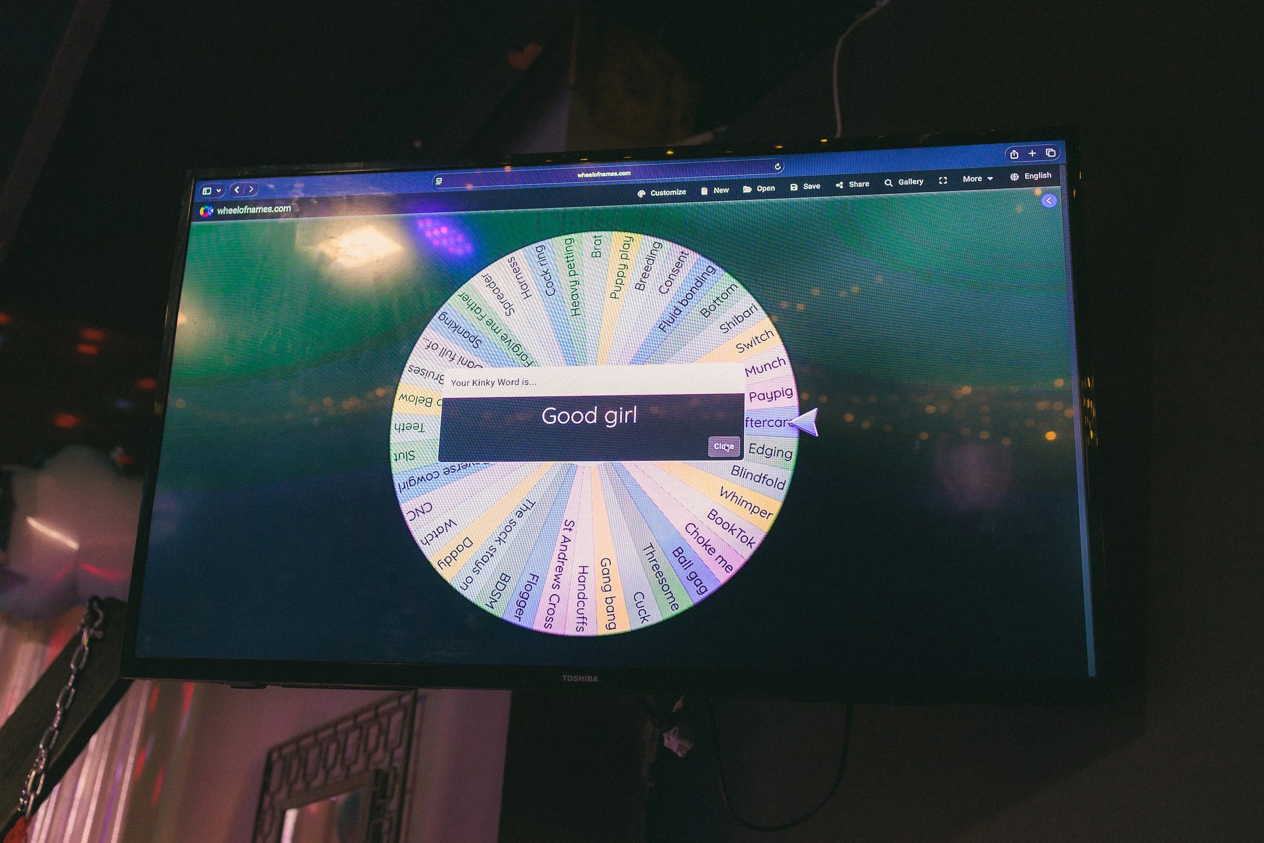 A computer monitor displays a spinning wheel labeled 'Your Kinky Word is...' with the phrase 'Good girl' inside.