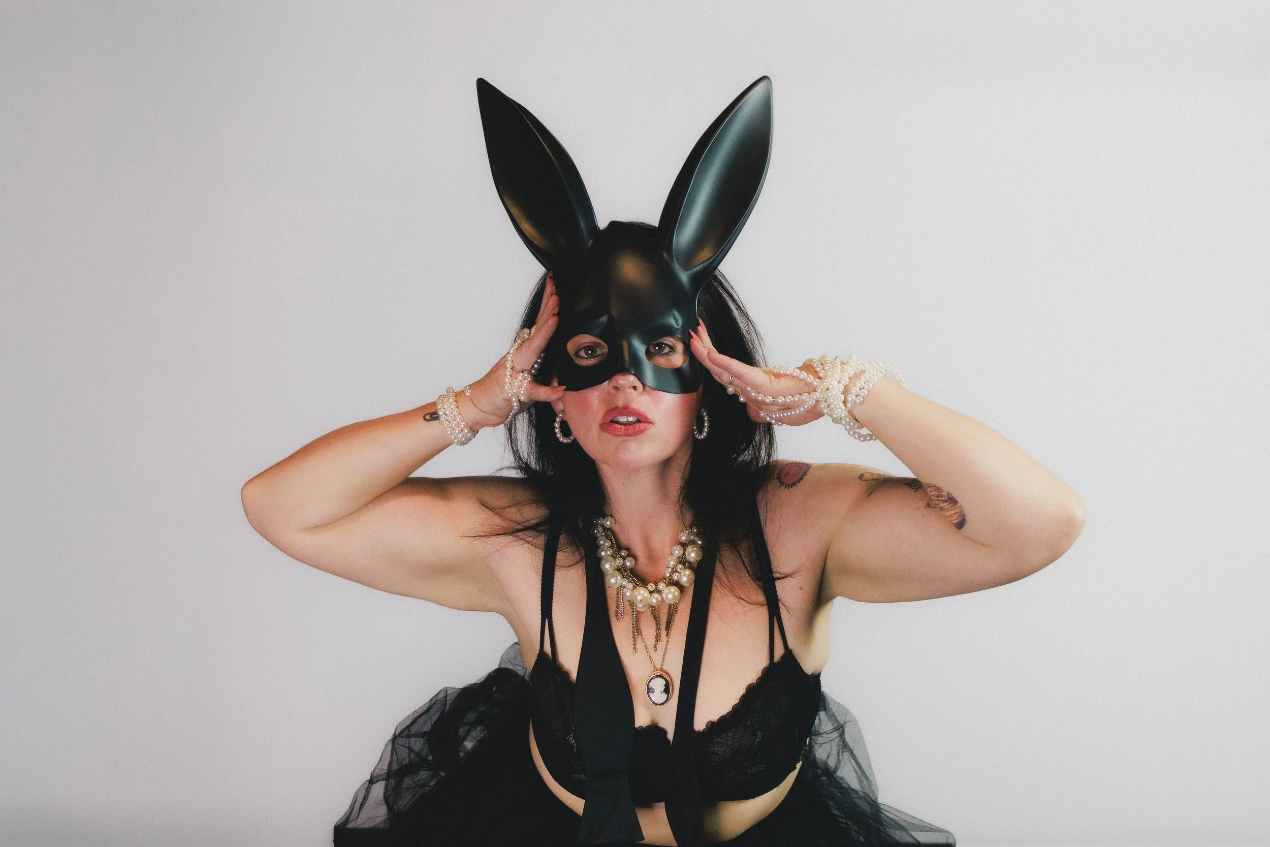 Woman wearing a black bunny mask, black lace lingerie, and pearl jewelry, posing against a plain white background.