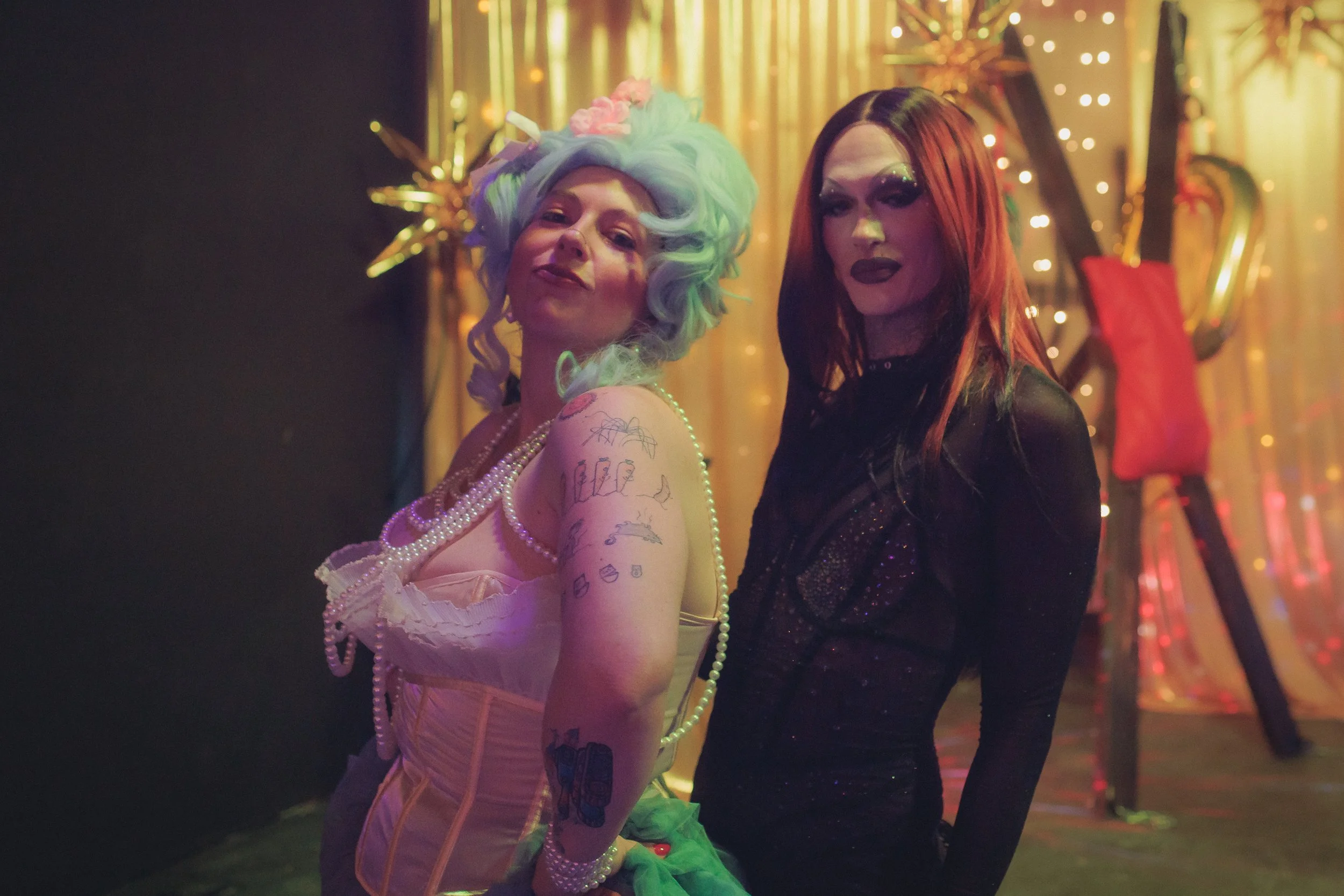 Two women dressed in vintage and dark gothic clothing, standing close together at a decorated event with string lights and balloons in the background.