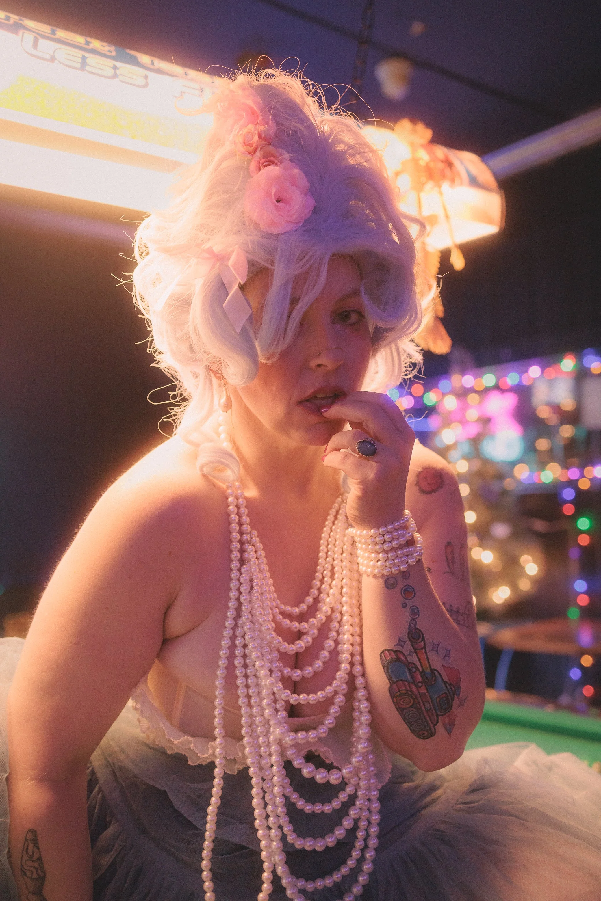 A woman with platinum blonde hair styled in vintage waves, wearing multiple strands of pearl necklaces, a pearl bracelet, and a large ring. She has tattoos on her arm and chest, including a colorful tattoo of a revolver and stars. The background is d