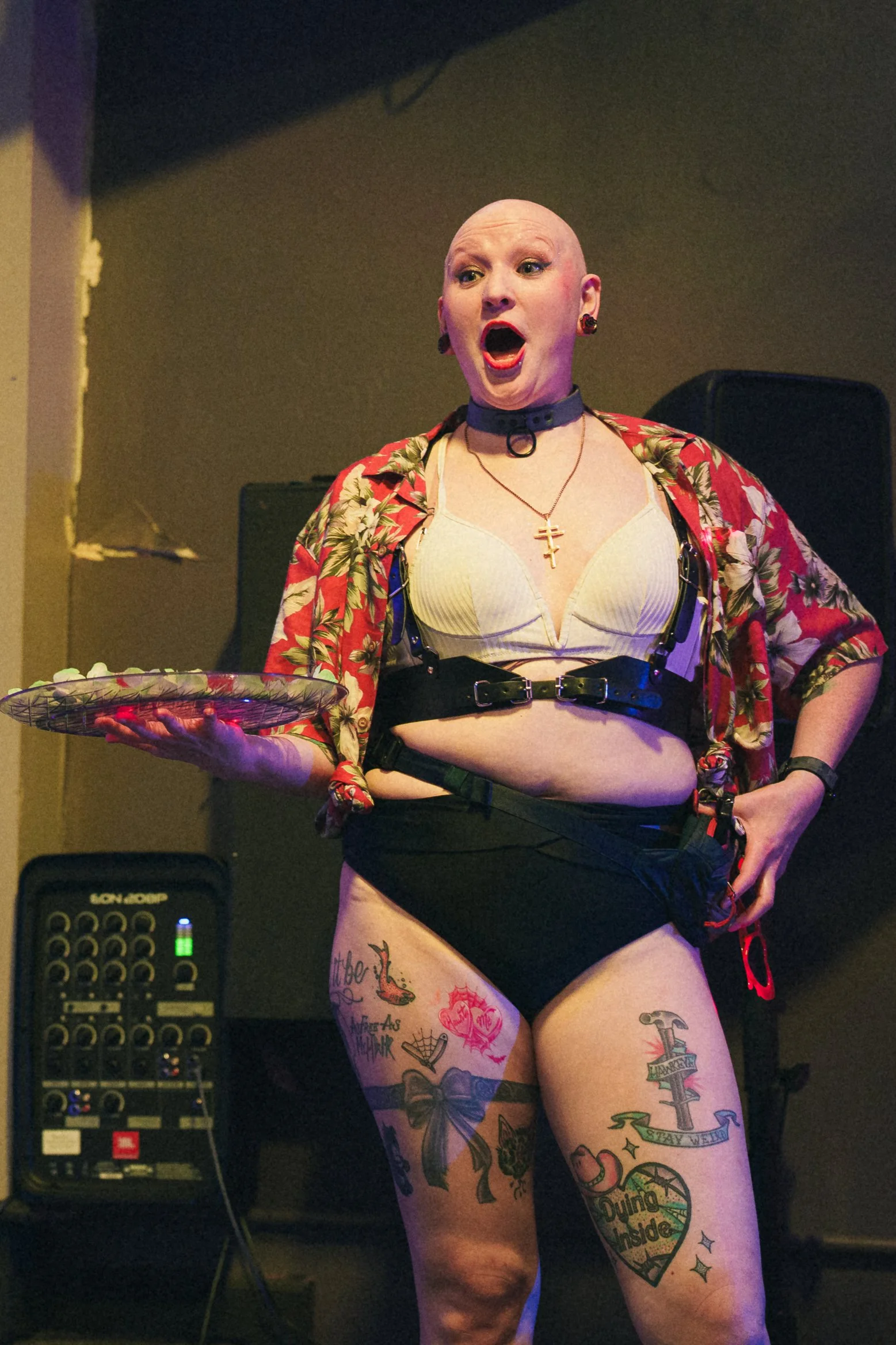 A person with a shaved head, colorful tattoos on legs, wearing a floral shirt, black lingerie, choker, and necklace, holding a tray, with sound equipment in the background and expressive mouth open.