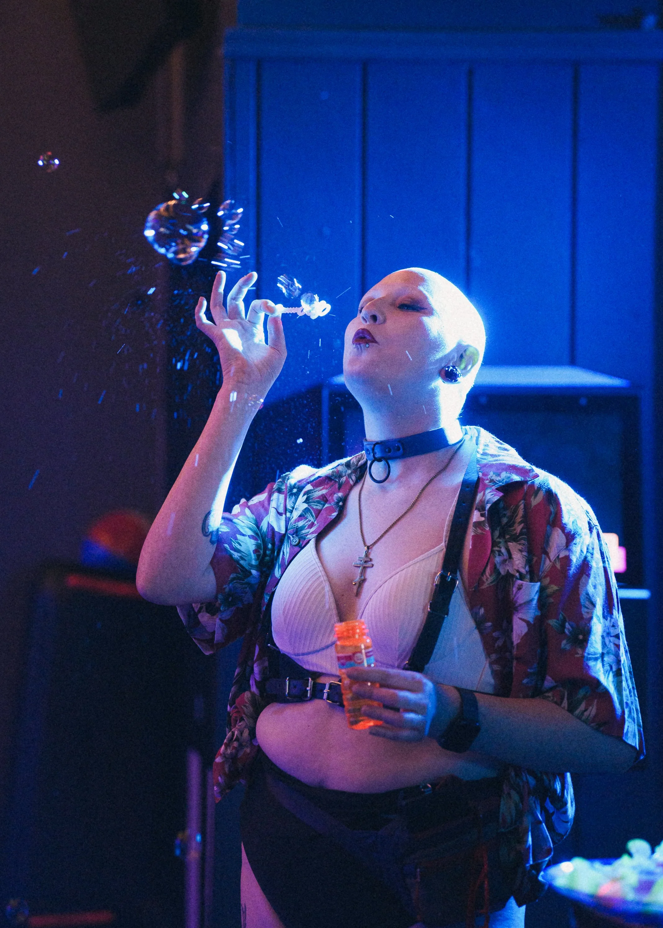 A woman with a shaved head, wearing makeup, earrings, and a choker, stands in a dimly lit room with blue and purple lighting. She is holding a small bottle of orange liquid in one hand and a lit cigarette in the other, with bubbles floating nearby.