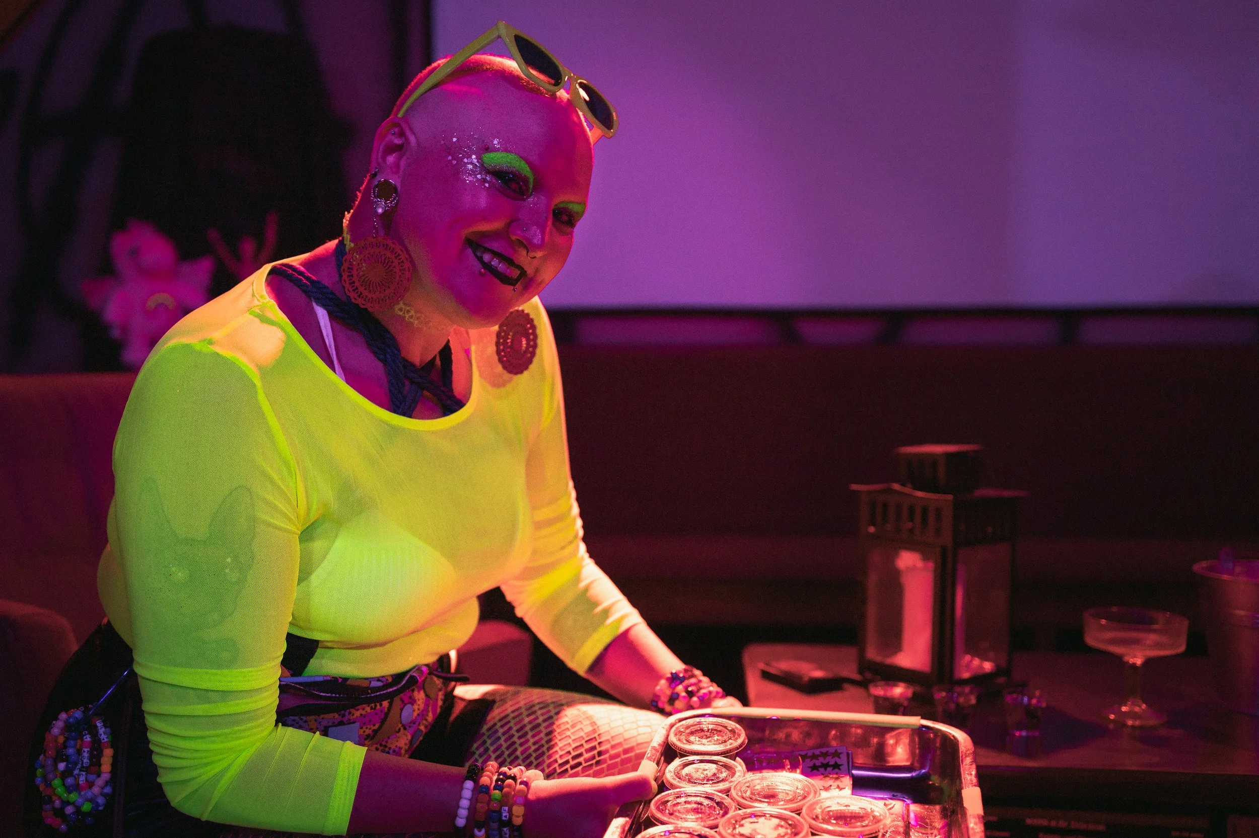 A person with colorful makeup and jewelry, wearing a neon yellow top and playing a DJ set in a dimly lit room with purple lighting.