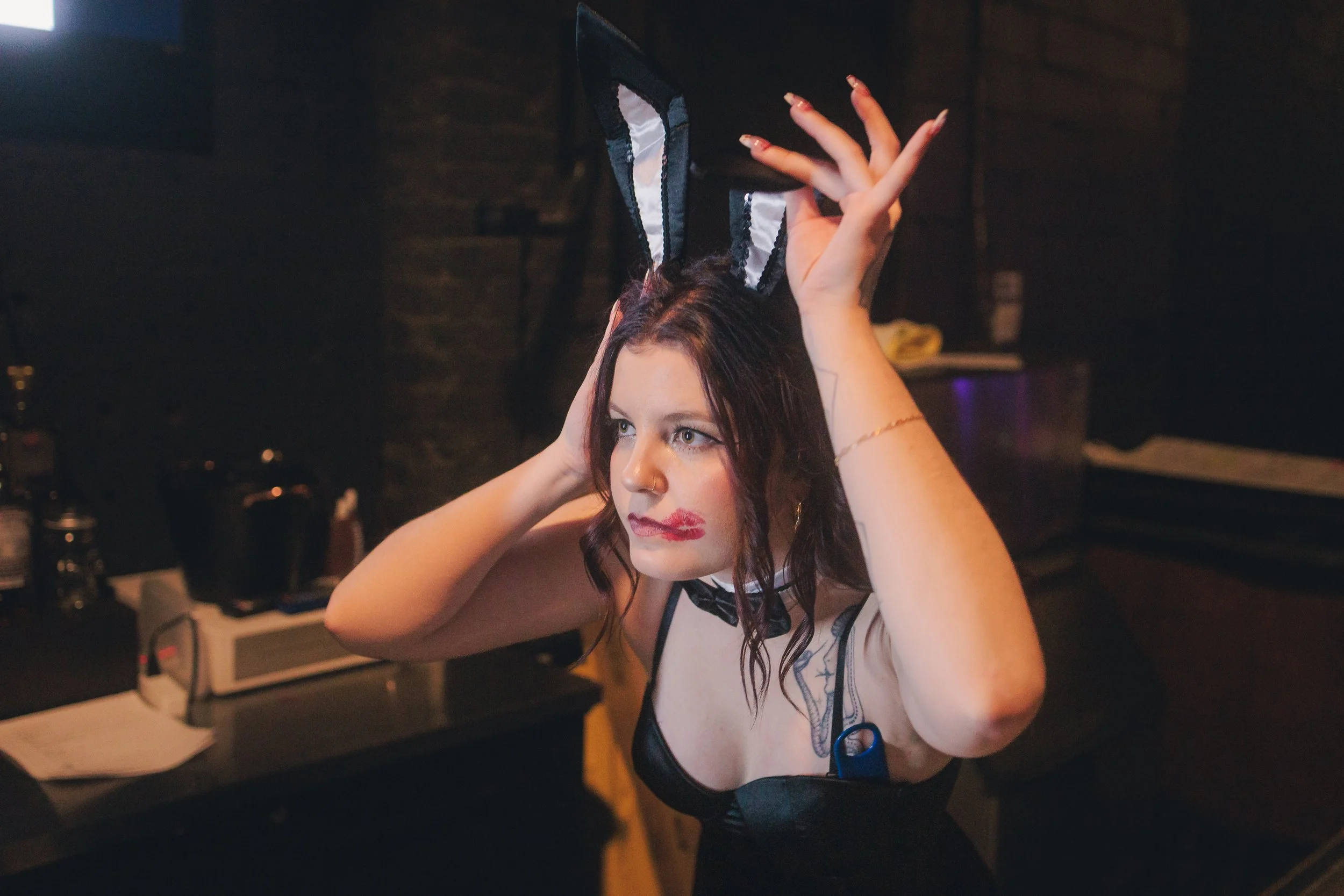 A woman with dark hair and makeup wearing bunny ears headband and a costume, adjusting the ears in a dimly lit room.