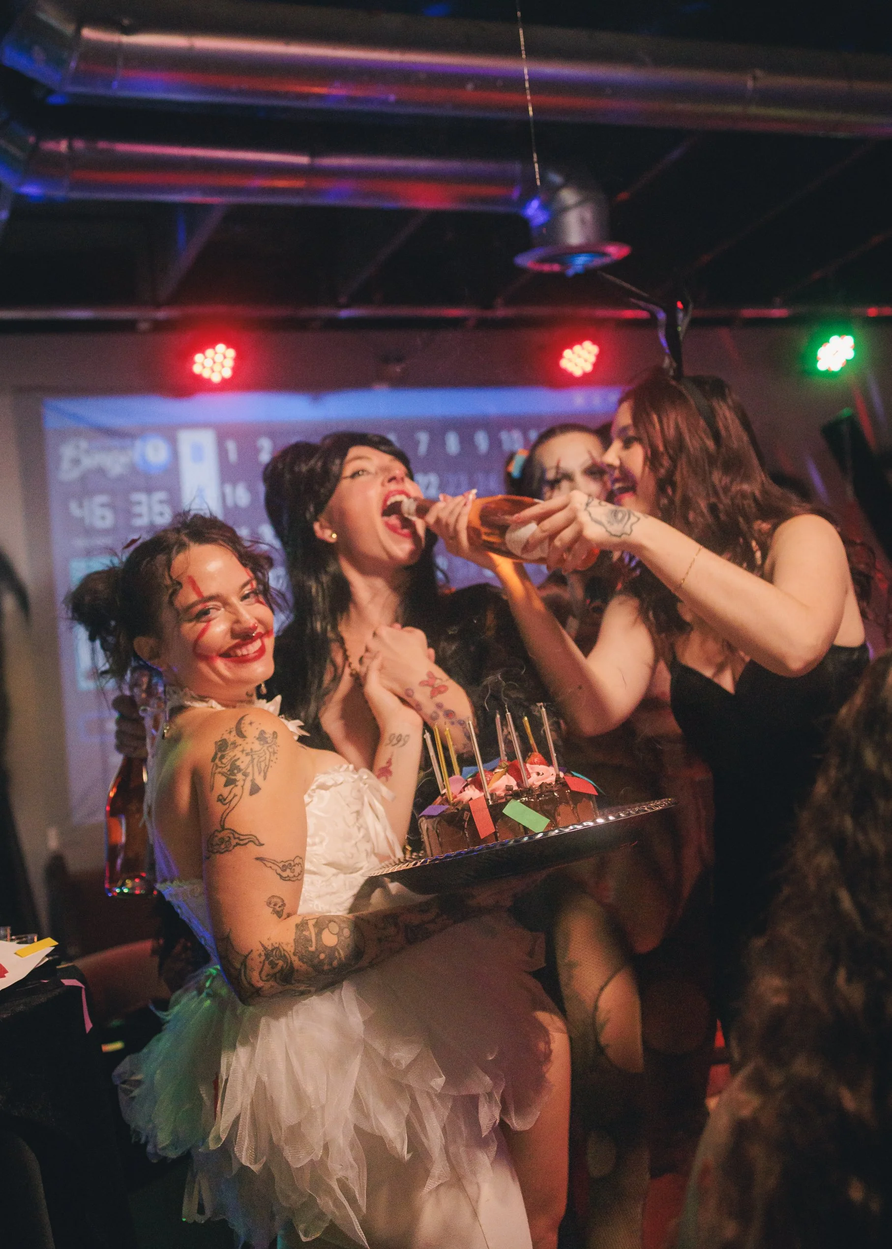 A group of women celebrating at a party, with one woman in a white dress holding a cake with candles, while others take a shot from a bottle. The scene is lively with colorful lighting and a background with polka dots and random objects.