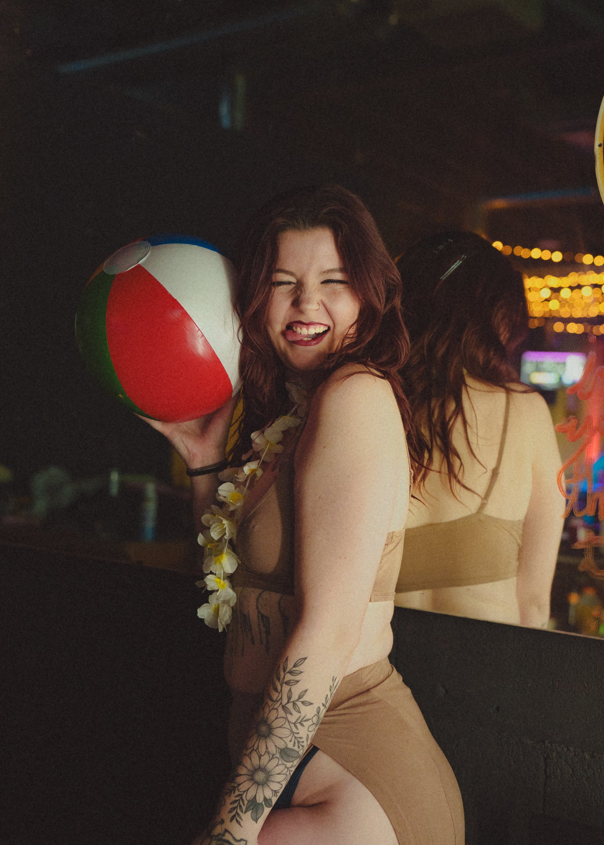 A woman with red hair smiling and holding a beach ball at a nighttime party. She is wearing a beige outfit and has floral tattoos on her arm. The background is blurred with colorful lights.
