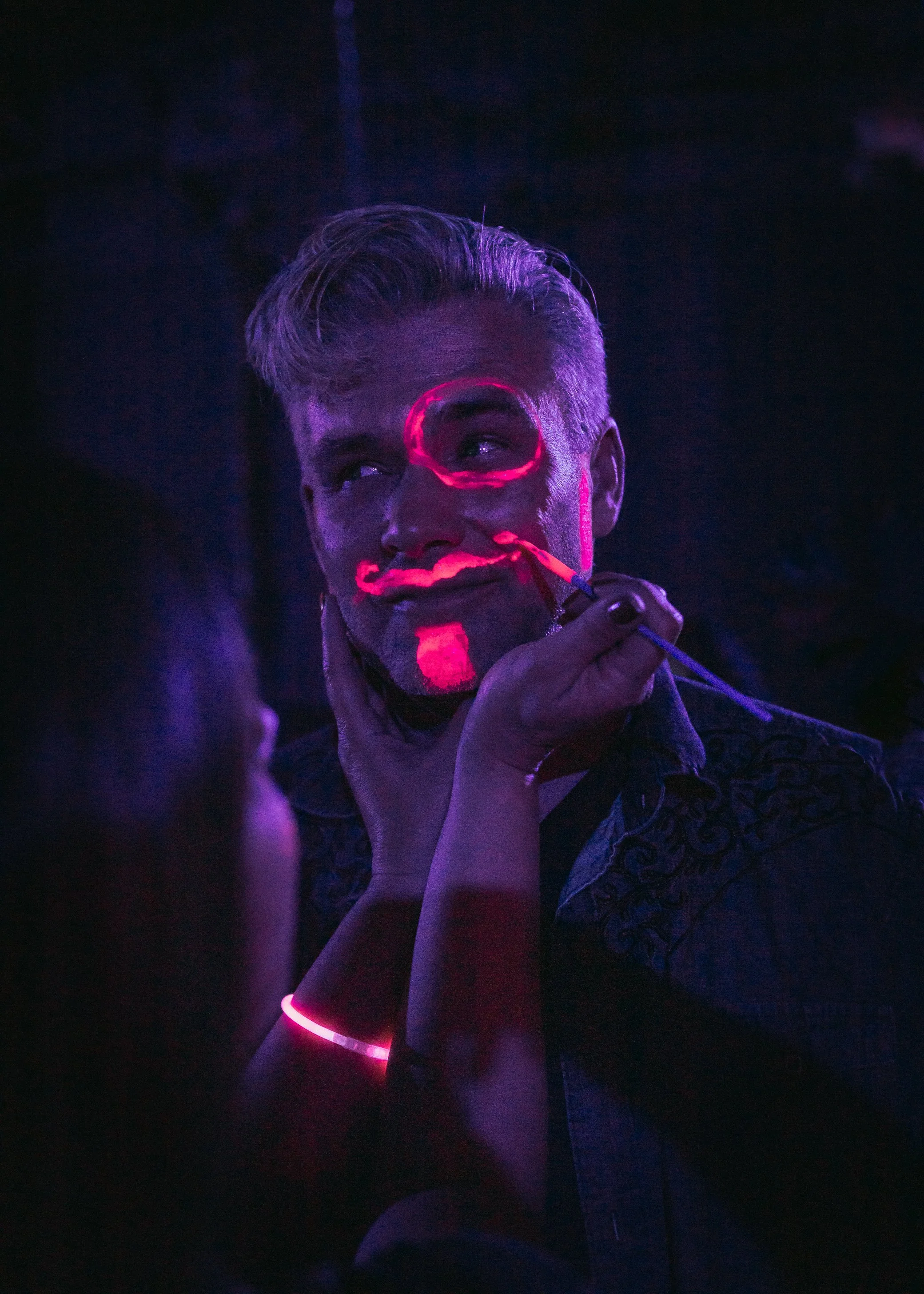 Person with a short hairstyle applying neon face paint under blacklight, wearing a glow bracelet, in a dark environment.