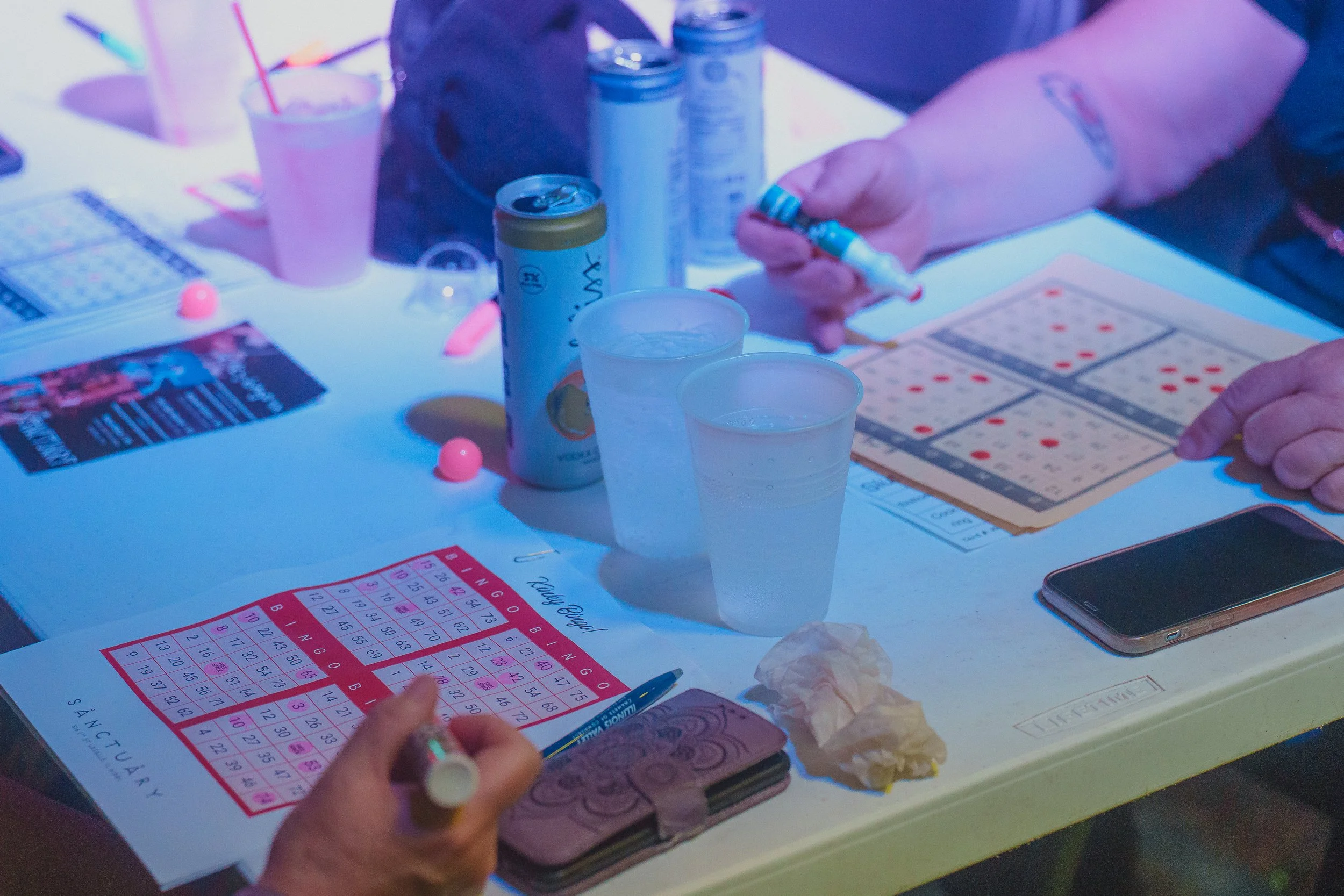 People playing bingo at a table with bingo cards, pills, drinks, and snacks.