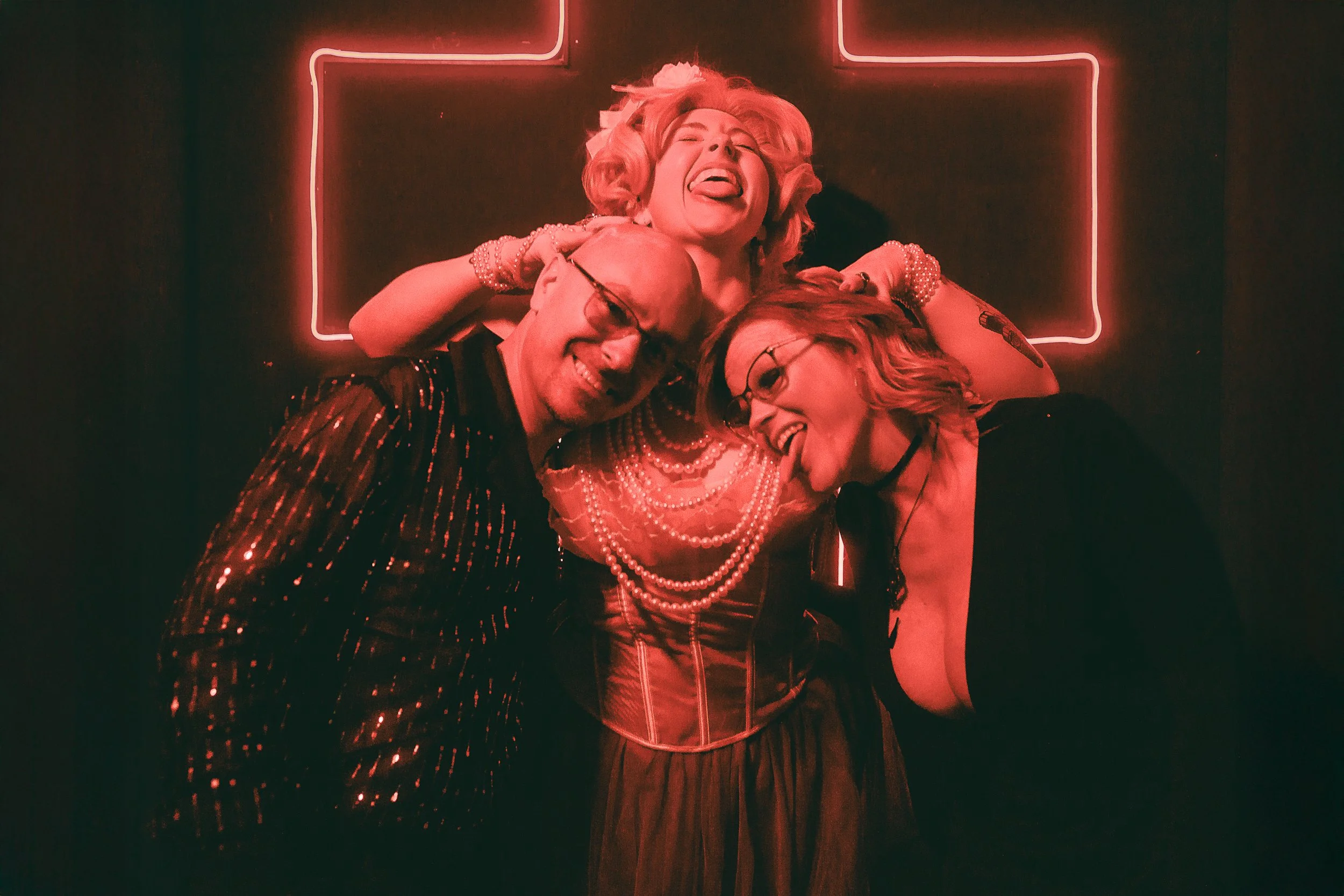 Three people joyfully hugging under a neon cross sign in a dark setting, with two women wearing glasses and a woman with blonde hair, all smiling and showing excitement.