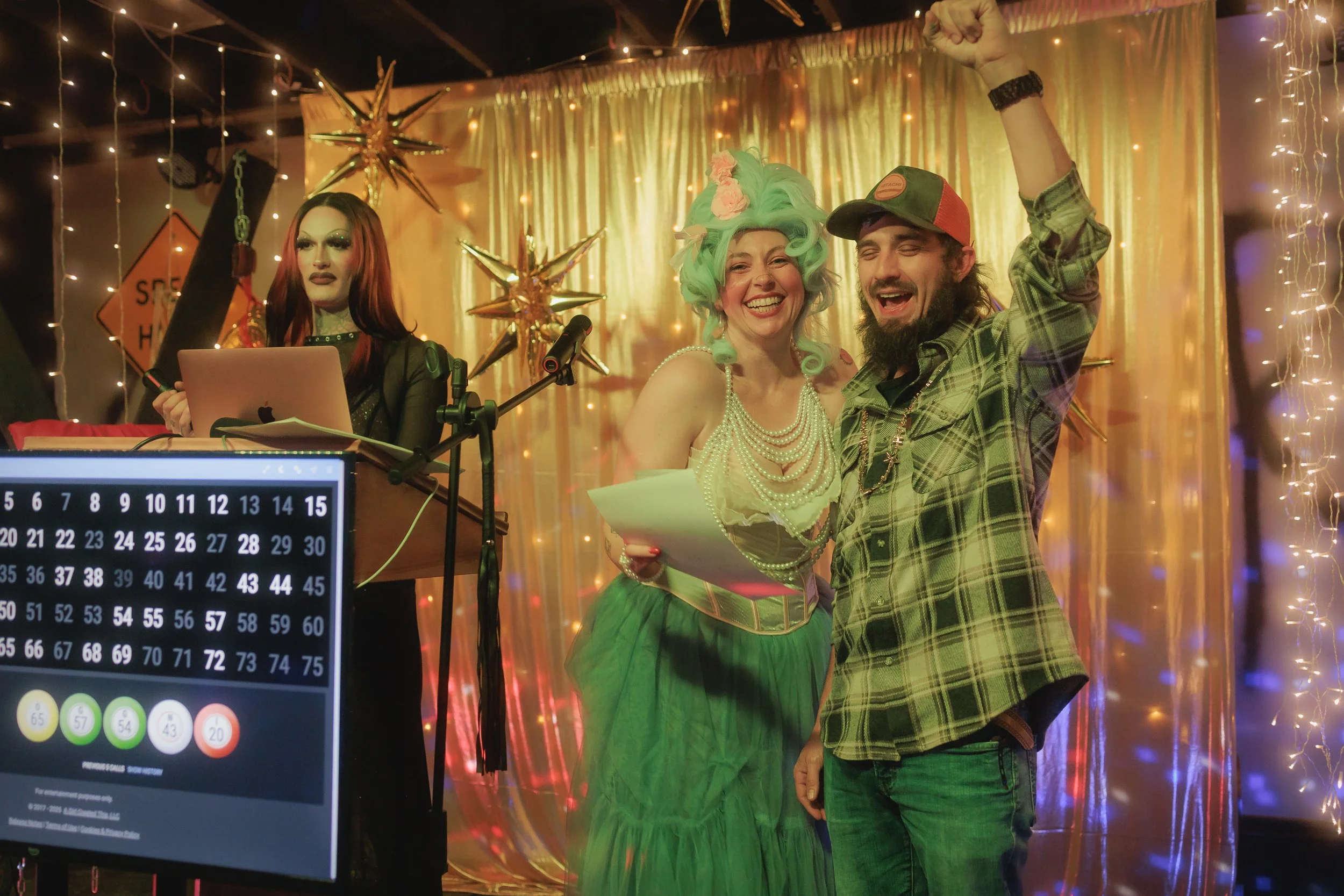 Happy couple celebrating, the woman with a green wig decorated with pink flowers and a pearl necklace, the man in a plaid shirt and trucker hat, raising his fist in the air. A woman with long red hair and dark makeup is seated at a table with a lapto