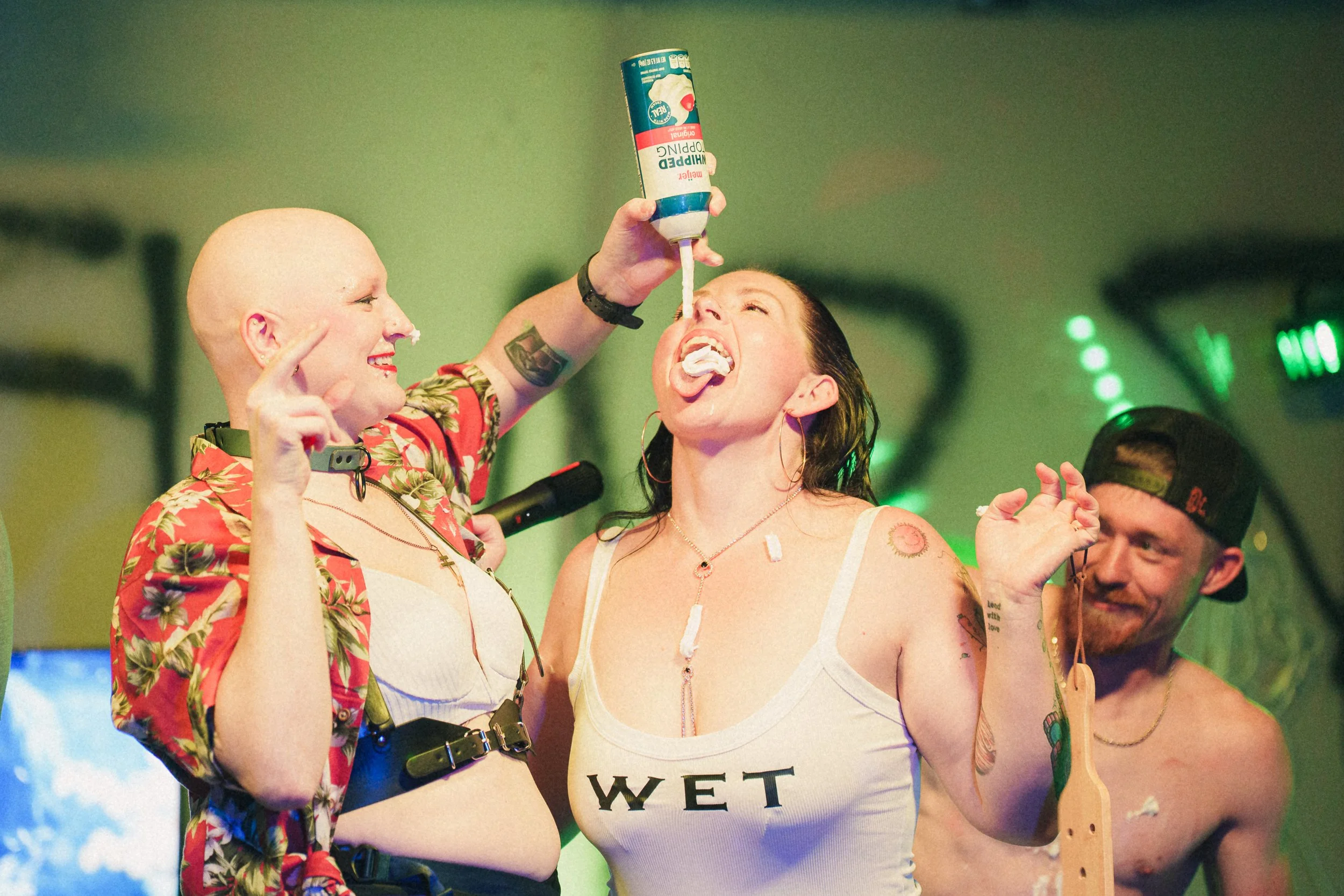 Two women and one man in a lively setting. The woman on the left is bald, smiling, and wearing a floral shirt, with some cream on her nose and pointing upward. The woman in the middle has dark hair, is sticking out her tongue, and is holding a can of