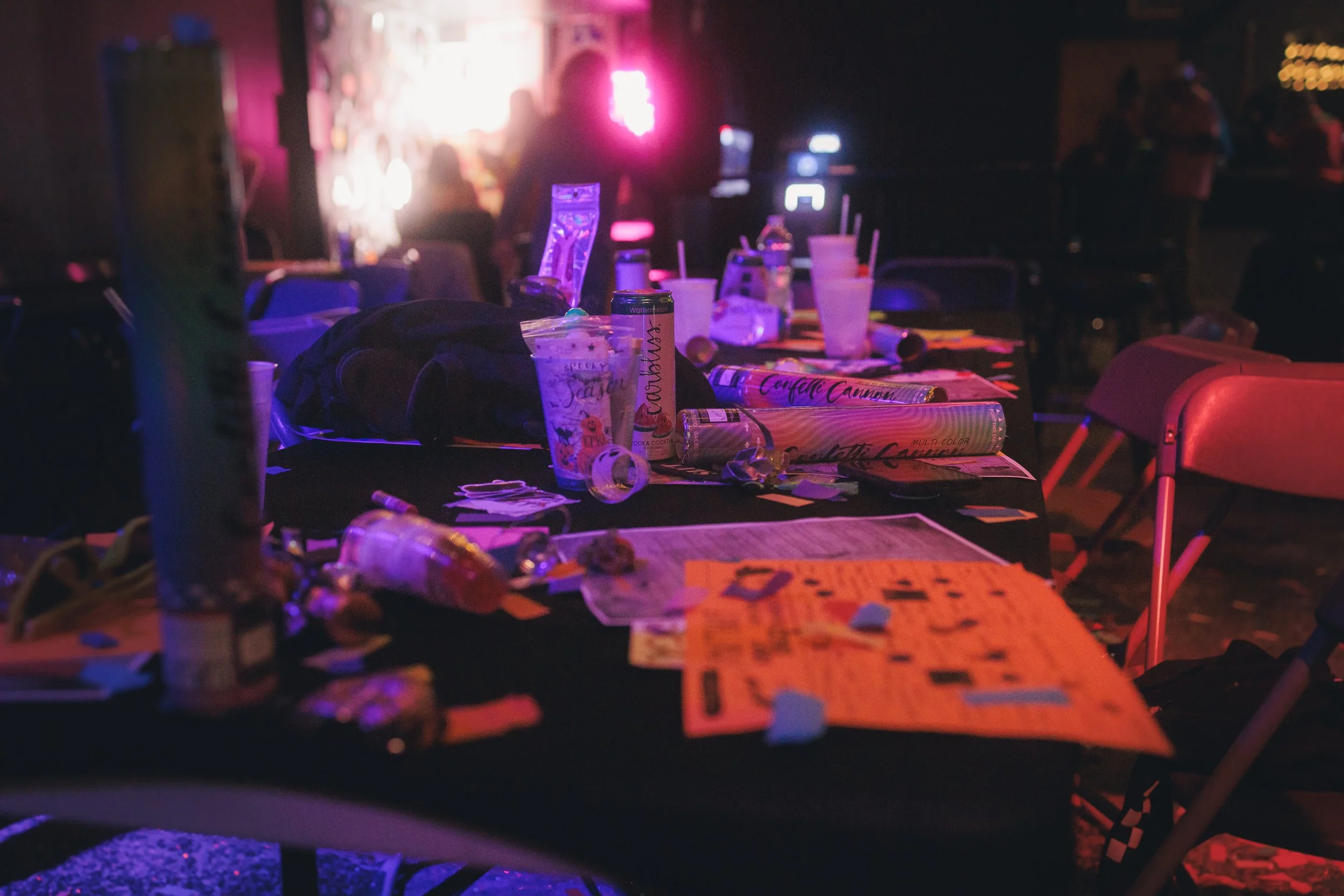 A table cluttered with various items, including snack containers, drinks, and scattered papers, in a dimly lit setting with colorful neon lights and silhouettes of people in the background.
