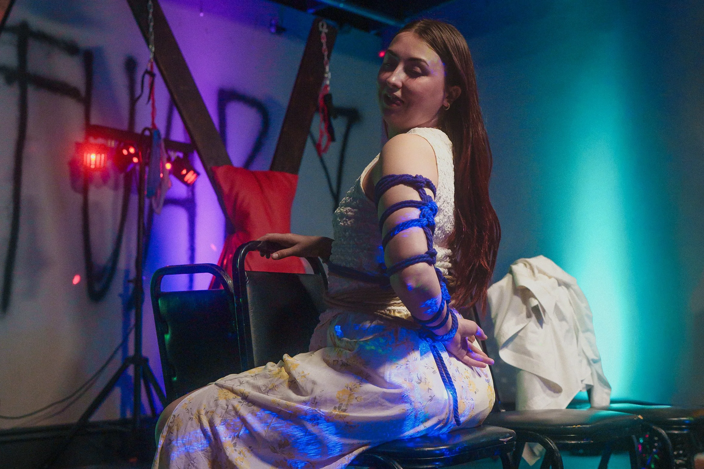 A woman with long red hair sitting in a dimly lit room with colorful lighting, surrounded by medical or scientific equipment, with her hands and arms bound with blue ropes, wearing a white dress with yellow floral patterns.