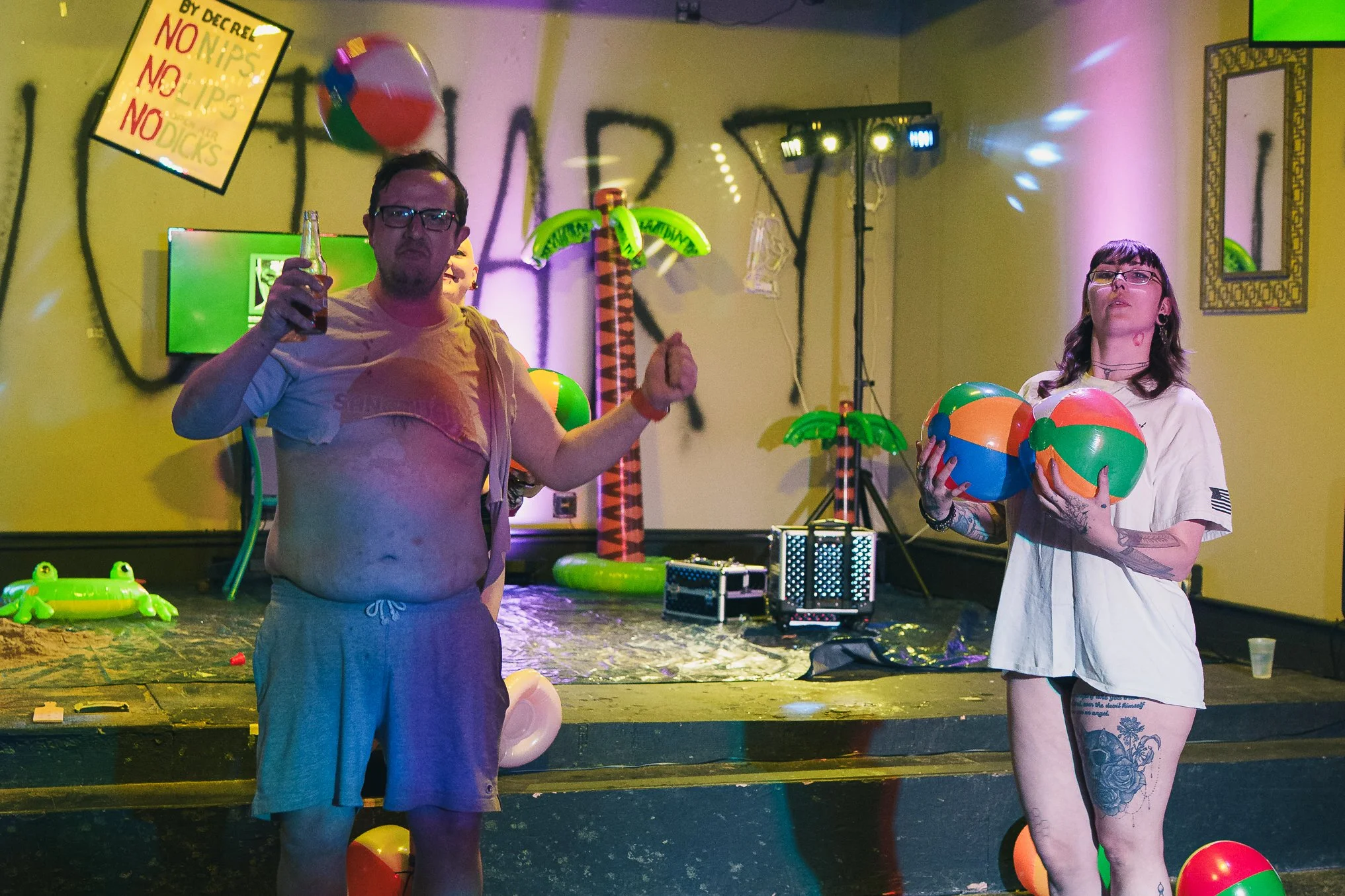 Two people standing on a stage decorated with colorful inflatable palm trees, beach balls, and a sign that reads 'NO DRINKS NO POLLEN'. The man on the left is shirtless, wearing glasses and khaki shorts, holding a bottle in his right hand and making 
