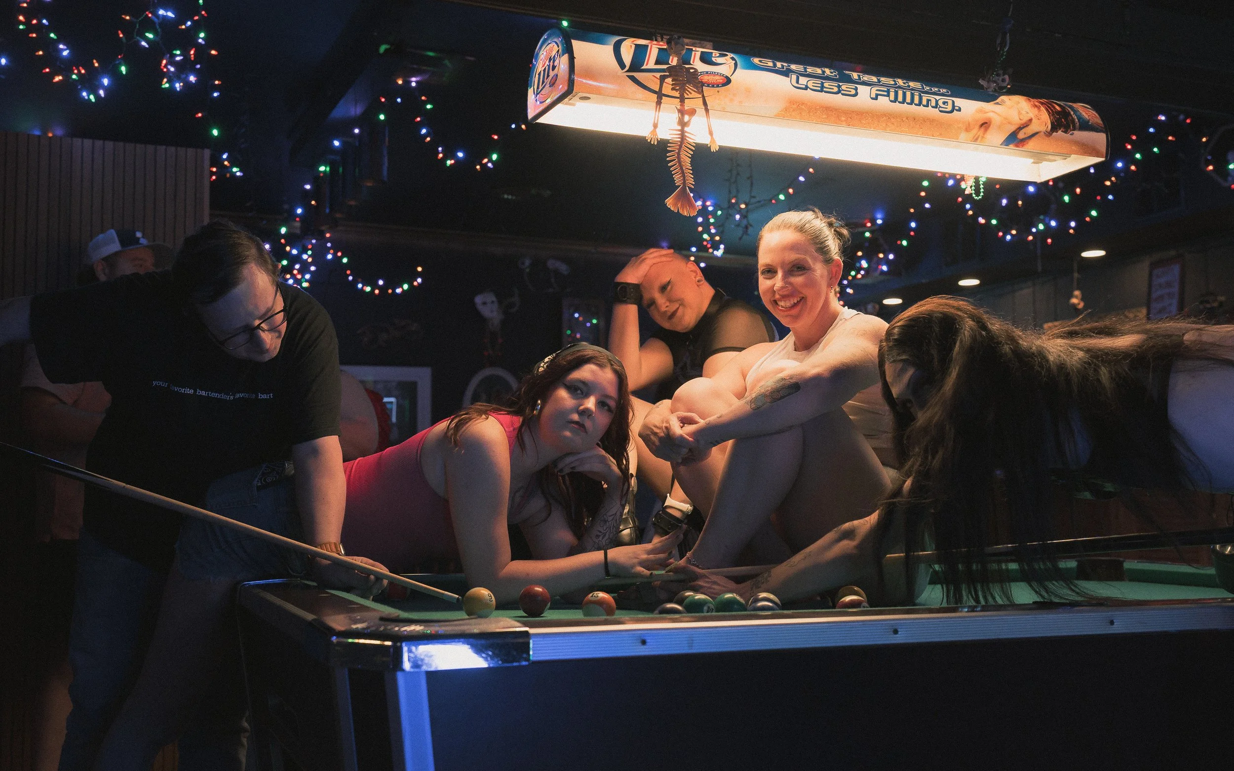 People playing pool in a bar decorated with colorful string lights, with some individuals leaning over the pool table and others standing around.