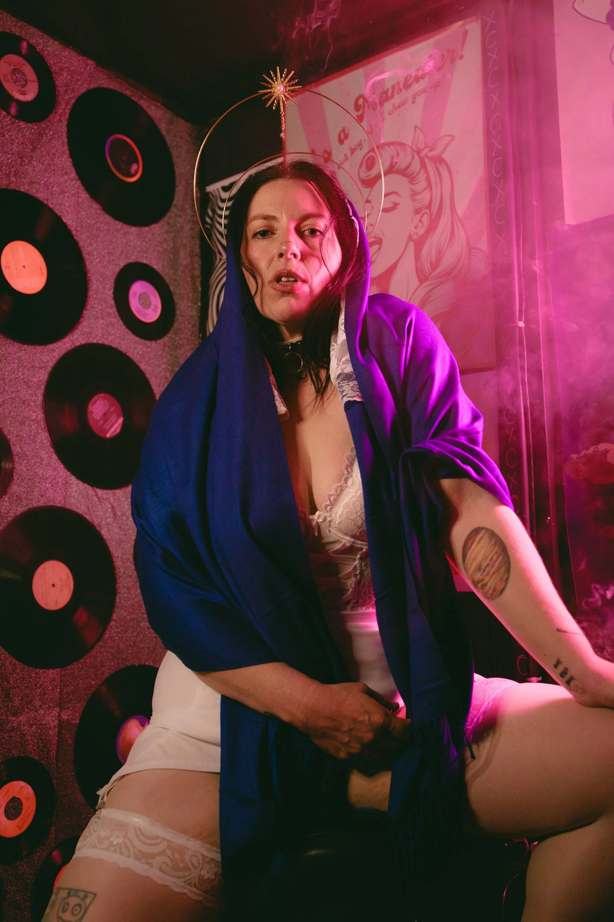 A woman with a halo headband, wearing a blue jacket and white lingerie, sitting on a man with tattoos. The background has privacy records and a portrait of a woman with hair styled in a vintage fashion on the wall.