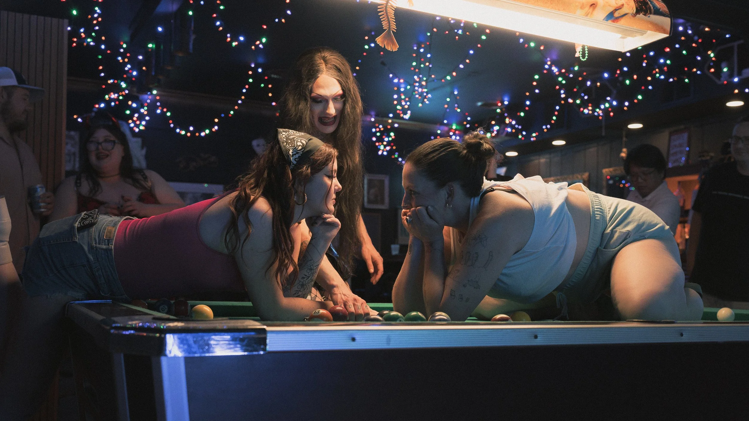 Three women playing pool at a bar, leaning over the table, with two of them supporting each other's heads. They are in a lively, festive environment decorated with colorful string lights hanging from the ceiling.