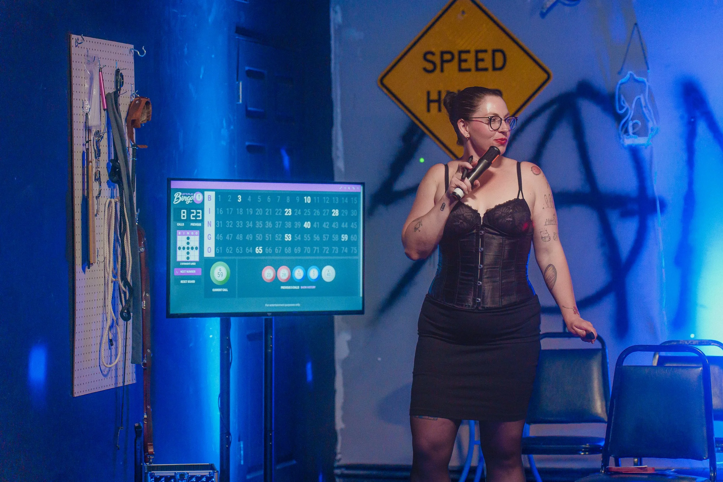A woman with tattoos and glasses holding a microphone stands in a room illuminated with blue lighting. Behind her, a computer screen displays a bingo game, and a pegboard on the wall holds various tools. A yellow sign reads 'Speed Hump' in the backgr