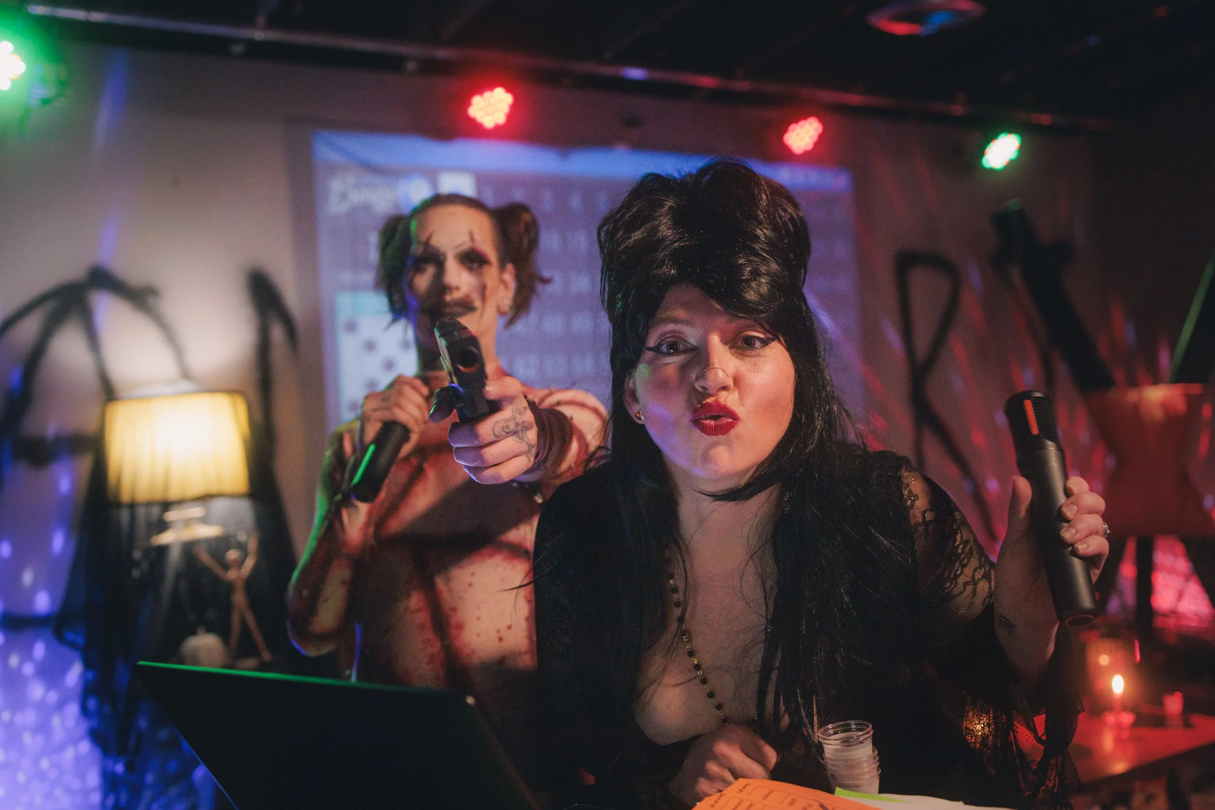 Two women are at a party or event, holding microphones and making playful expressions. One woman in the foreground has dark hair styled in a high beehive, wearing lipstick and a black lace top. The woman in the background has light brown hair in pigt