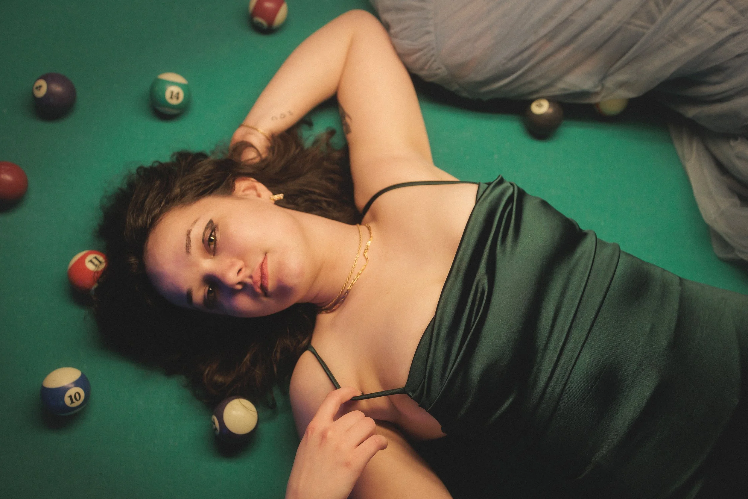 A young woman with dark wavy hair lying on a pool table among billiard balls, wearing a black satin dress and gold jewelry, gazing at the camera