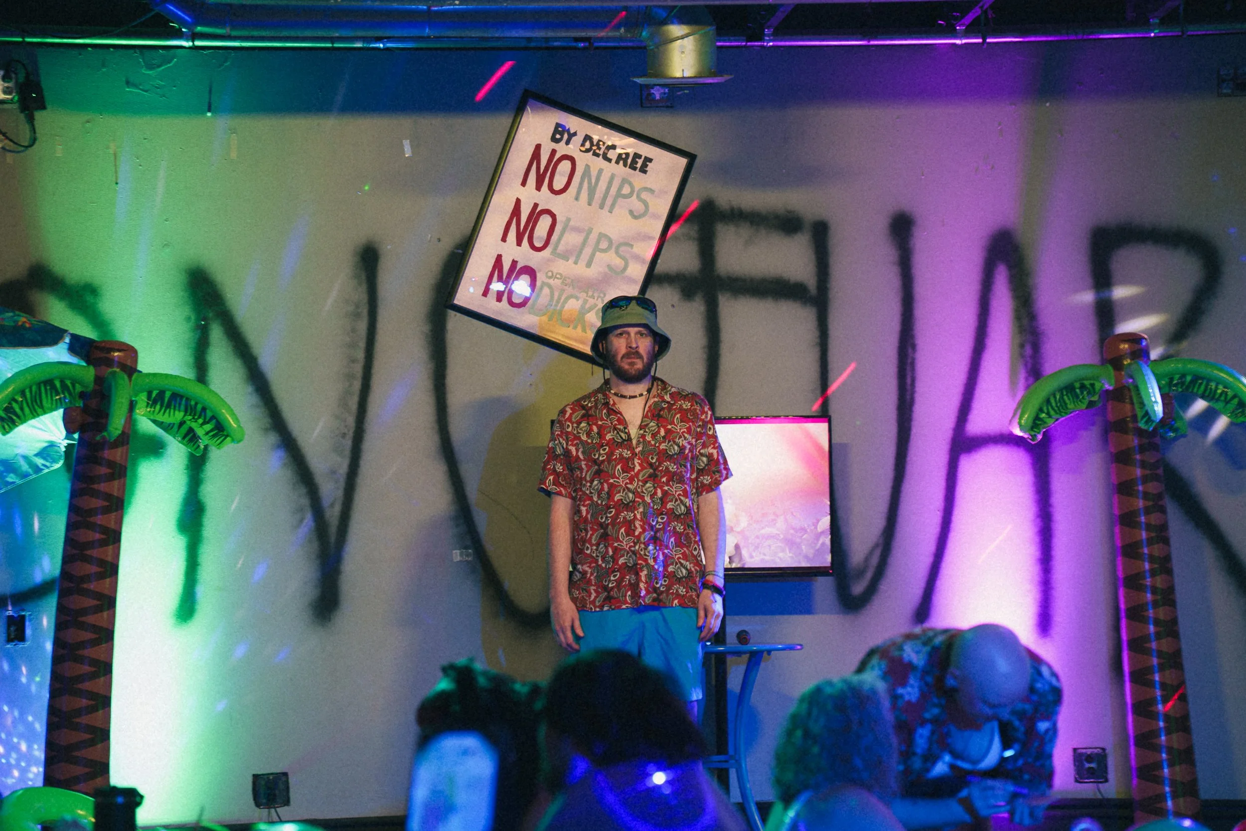 A man with a beard wearing a patterned shirt, shorts, and a bucket hat standing in a colorful, neon-lit room with inflatable palm trees, a sign that reads 'BY DECREE NO NIPS NO LIPS NO DICK,' and a crowd of people in the foreground.