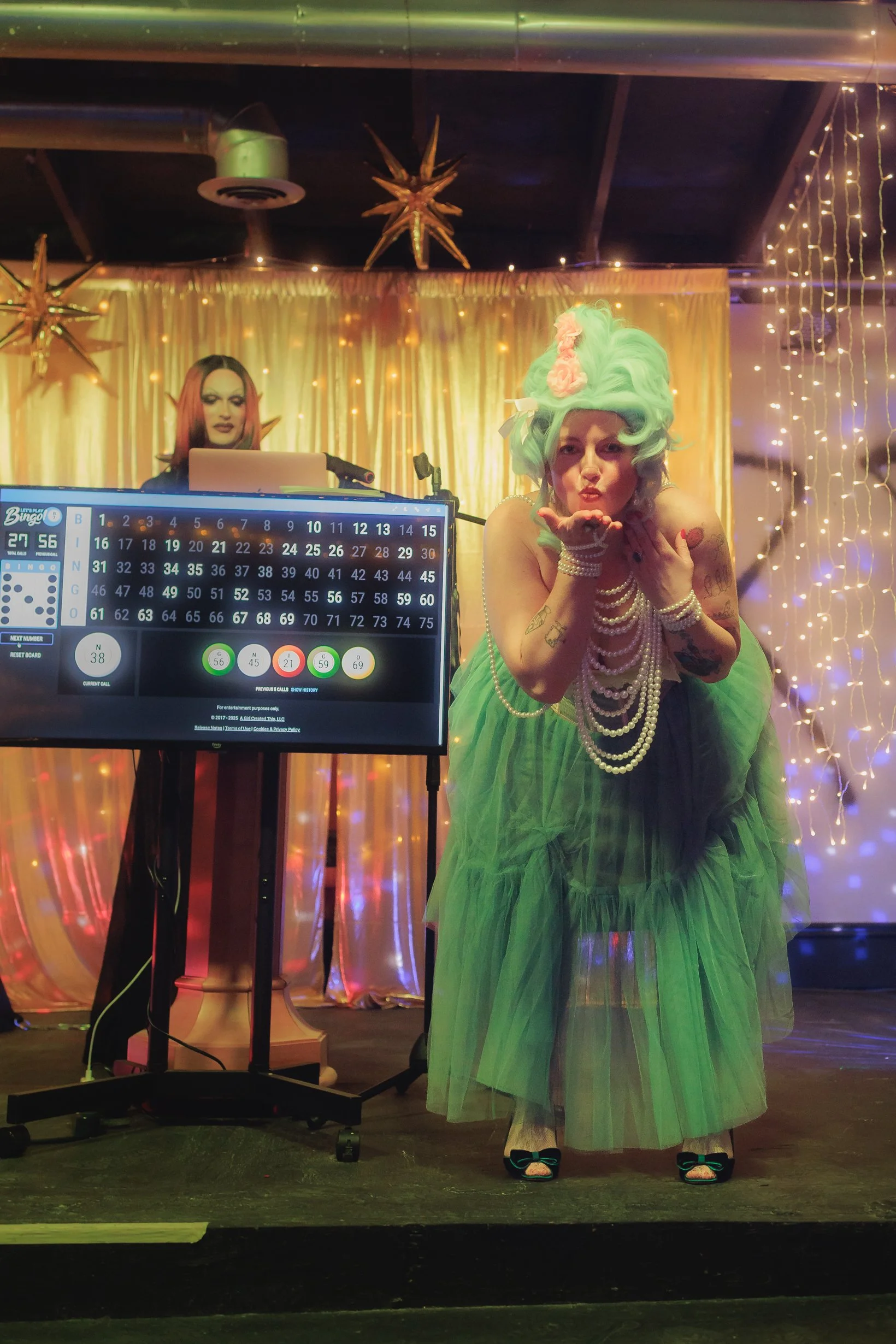 Person dressed in a green tulle dress with pearl jewelry and a mint green wig blowing a kiss at a bingo game, with a star-shaped decoration and string lights in the background.