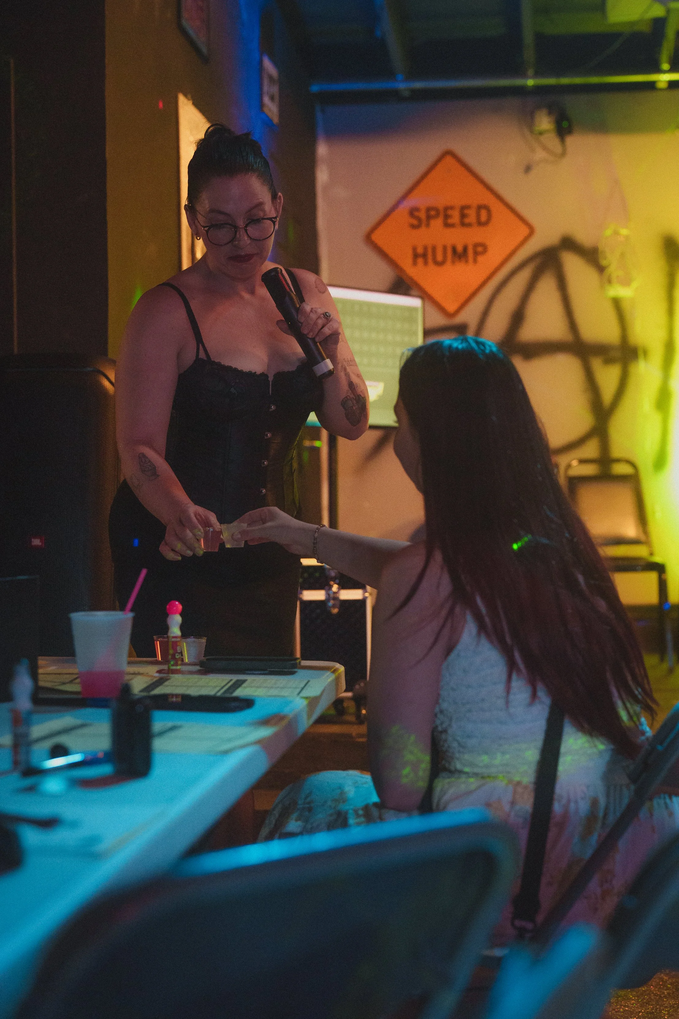 A woman with dark hair in a bun, wearing glasses and a black lace top, holding a microphone. She is engaging with a person sitting at a table in a dimly lit room with colorful neon lights and a sign on the wall that says 'Speed Hump.' The setting app