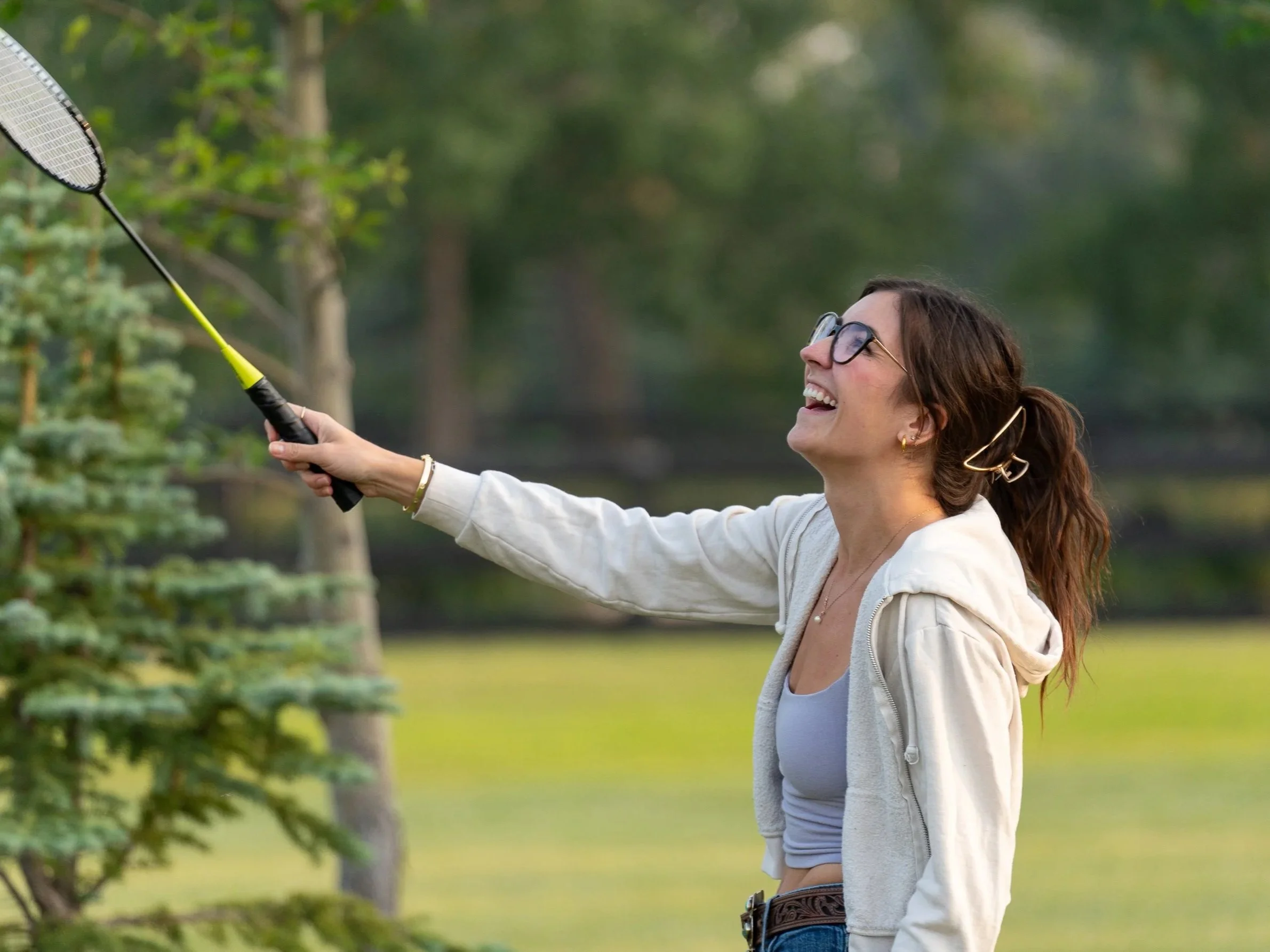 Woman with sunglasses playing badminton outdoors, smiling and holding a racket, surrounded by trees and greenery.