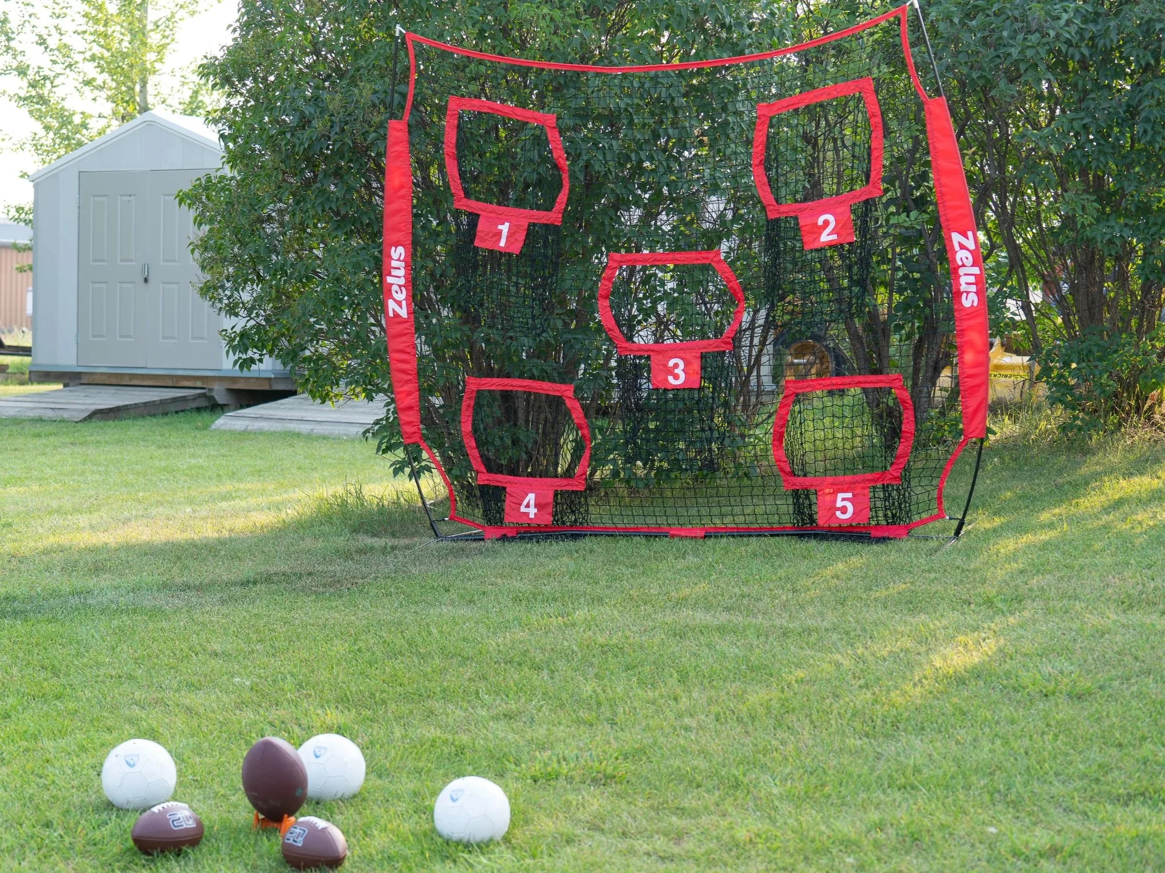 A backyard game set up with a black and red target net for football throws, numbered from 1 to 5, with multiple footballs on the grass in front.