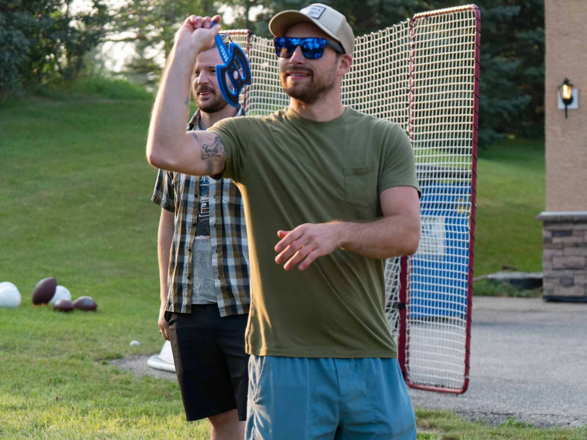 A man wearing sunglasses, a baseball cap, and a green T-shirt holds up a blue plastic object, smiling, with another man in a plaid shirt in the background. They are outdoors on a grassy area near a basketball hoop and some footballs.