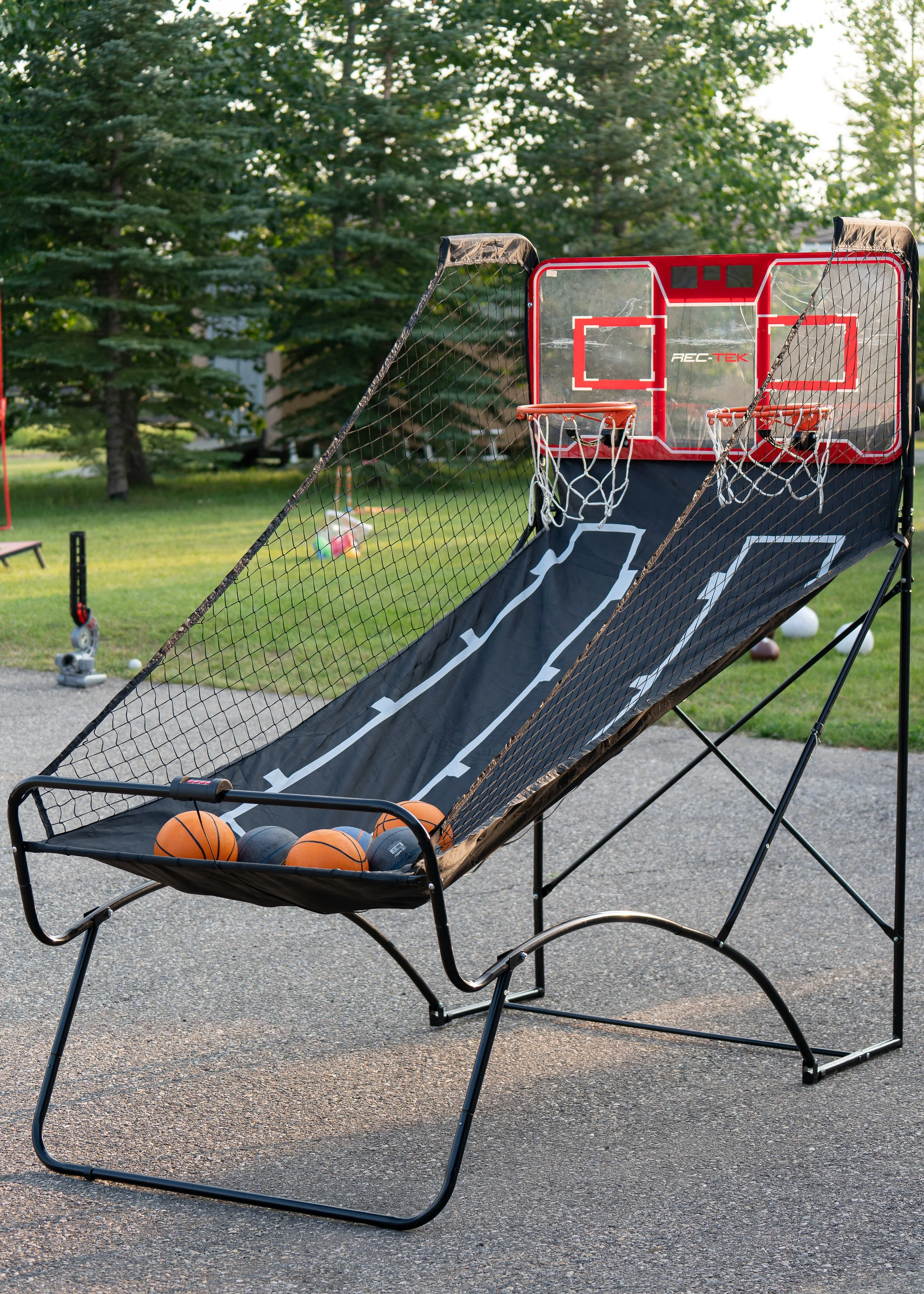 Outdoor basketball arcade game with basketballs in the tray, set up on a paved surface with trees in the background.