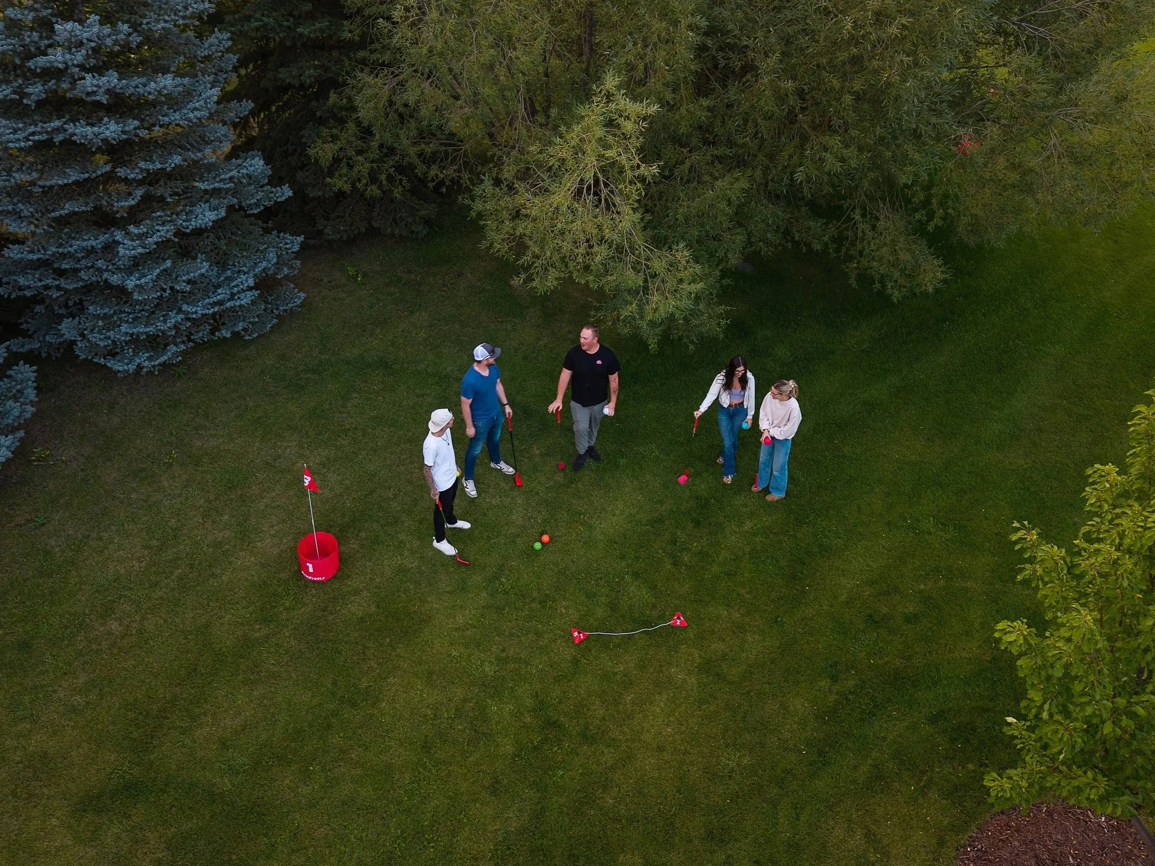 A group of six people playing mini-golf on a green lawn, surrounded by trees, with a red bucket and flags nearby.