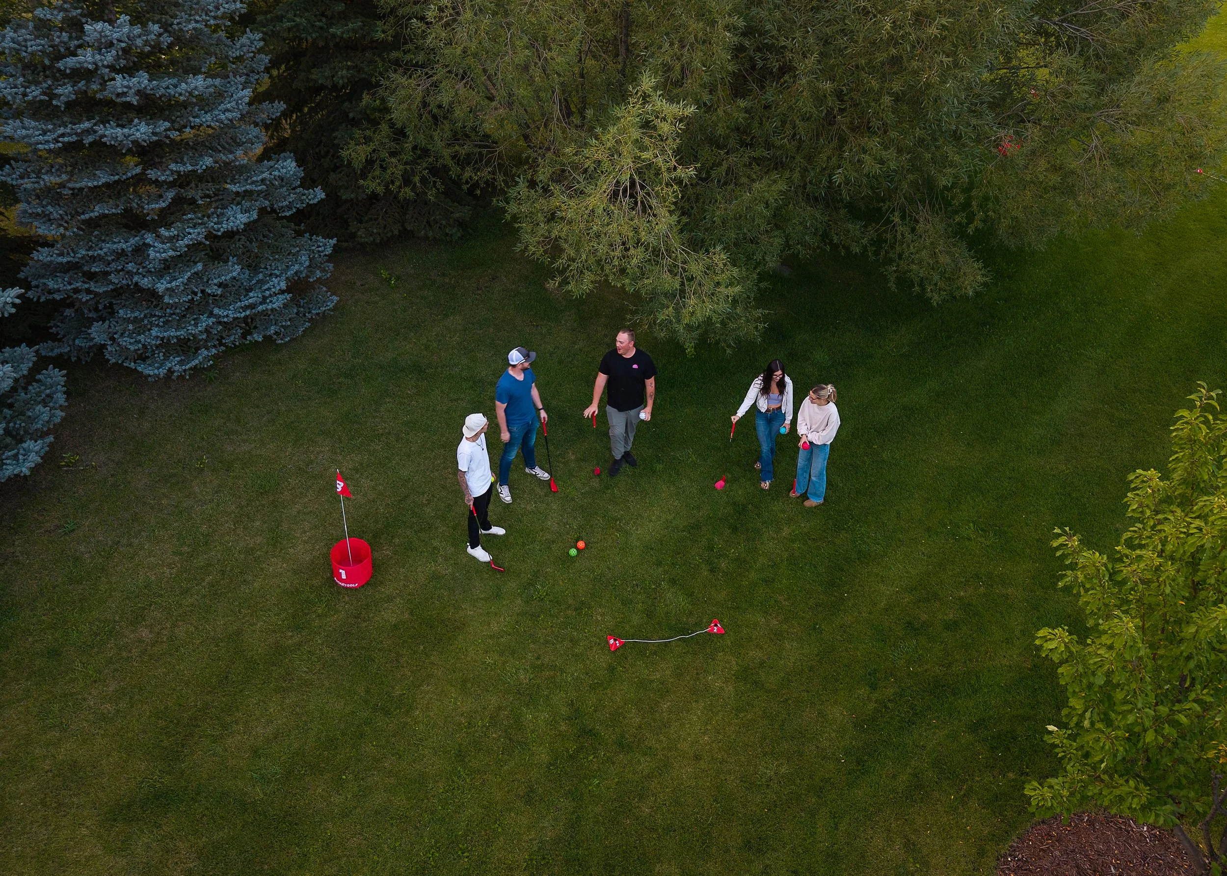 A group of five people playing golf on a grassy area surrounded by trees. They are holding golf clubs and standing near golf balls, with some equipment and flags visible.
