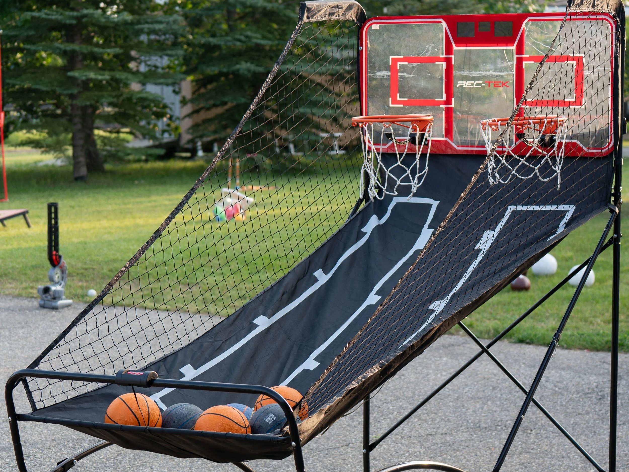 Basketball arcade game with basketballs inside, set outdoors on a paved surface with grass and trees in the background.