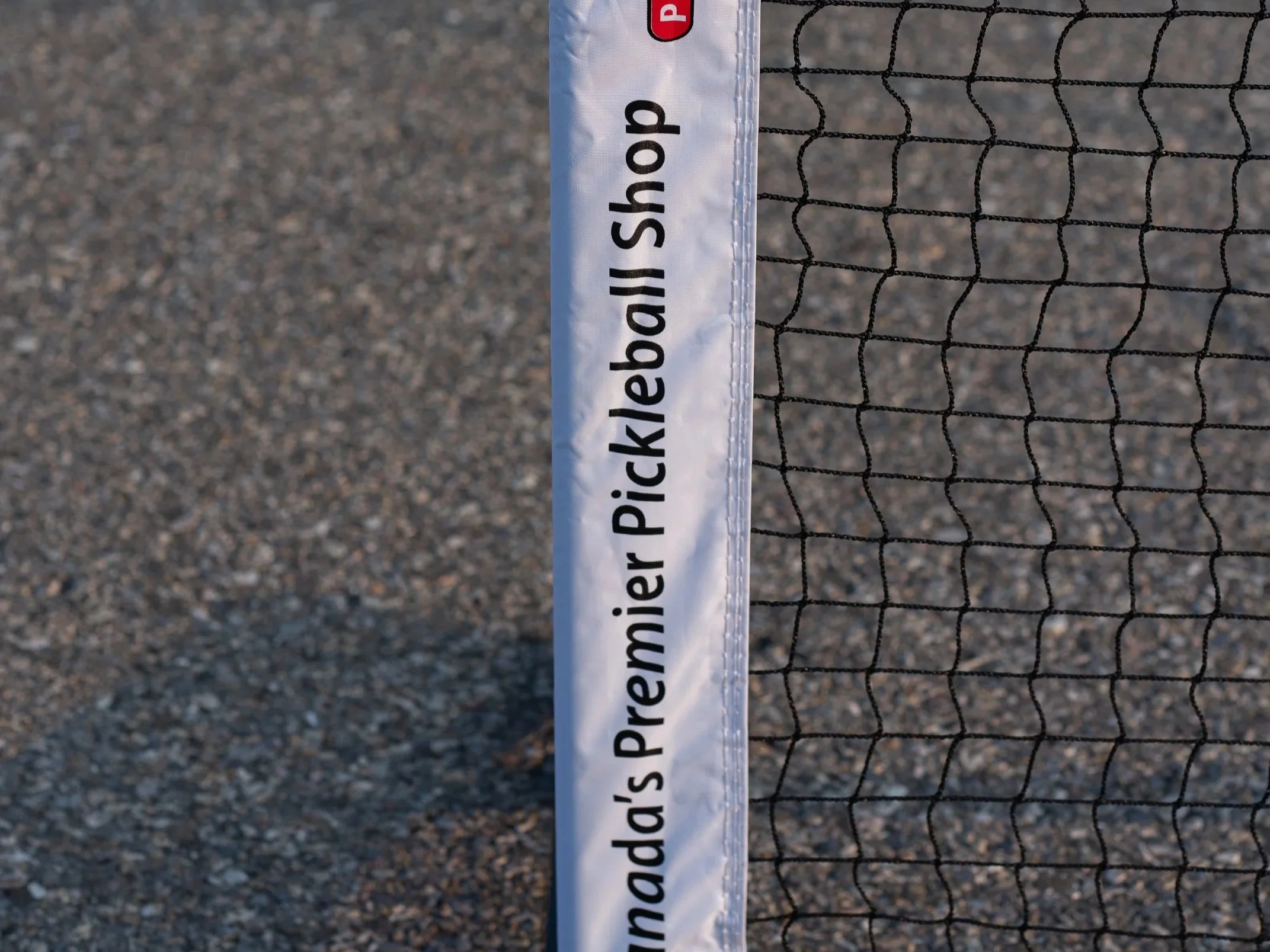 Close-up of a volleyball net and a pole with a label that reads 'Manda's Premier Pickleball Setup' on a gravel surface.