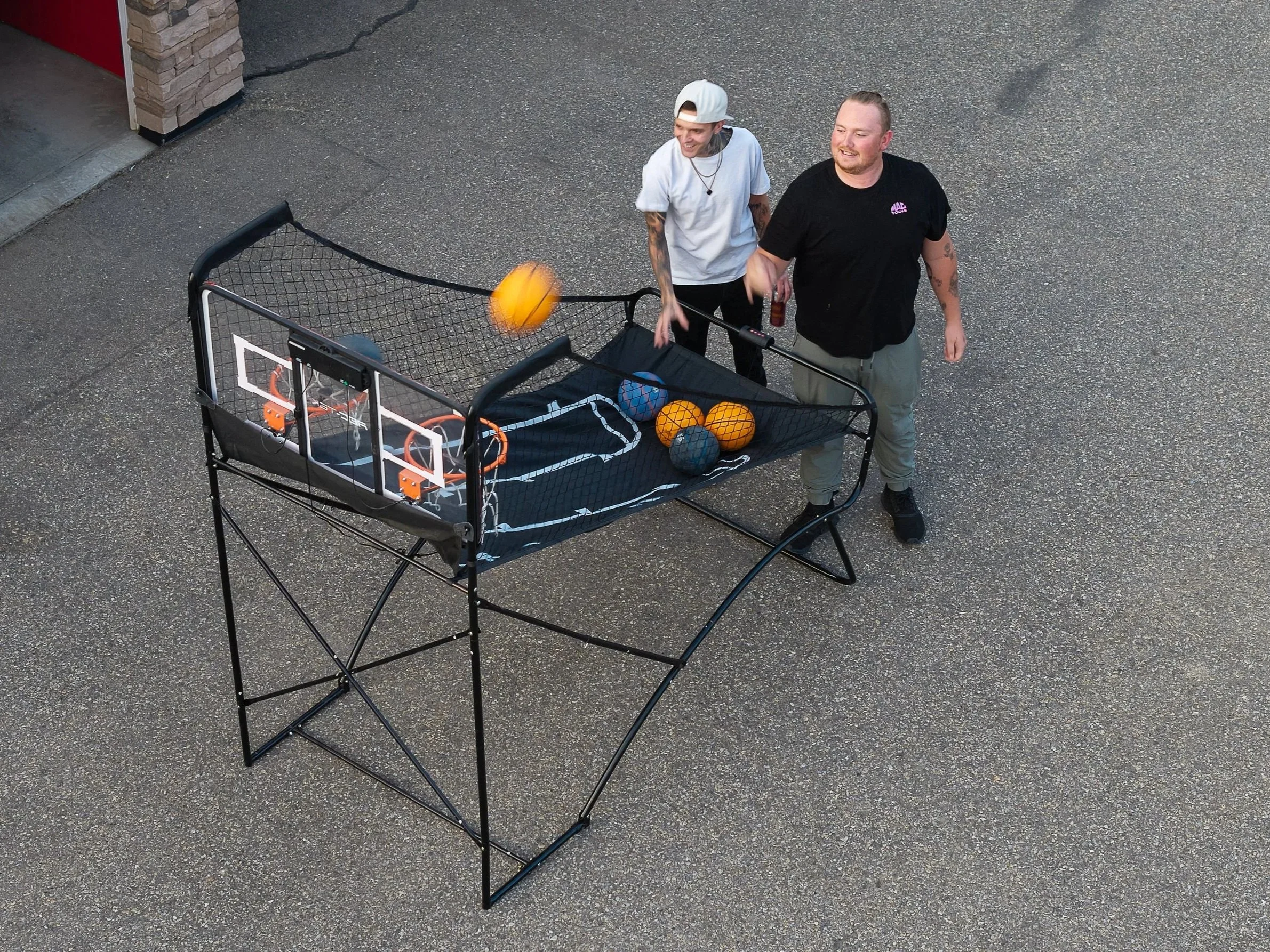 Two men playing basketball in a backyard with a mini basketball hoop and a ball return device.