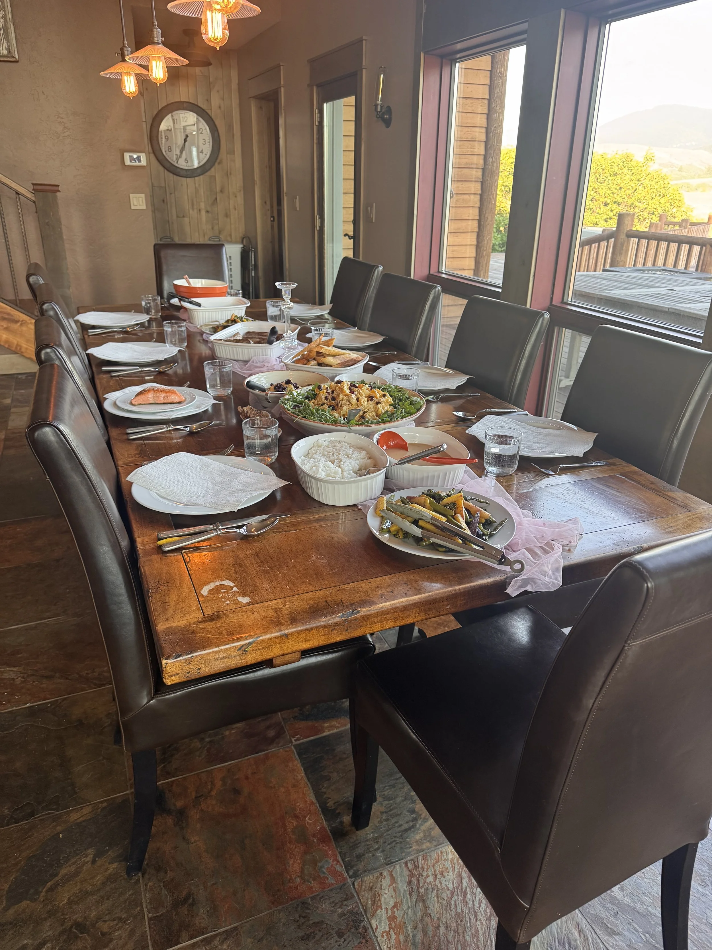A dining table set for a meal with various plates of food, glasses, and utensils, in a room with large windows that show an outdoor view of trees and a deck, decorated with hanging pendant lights and a wall clock.