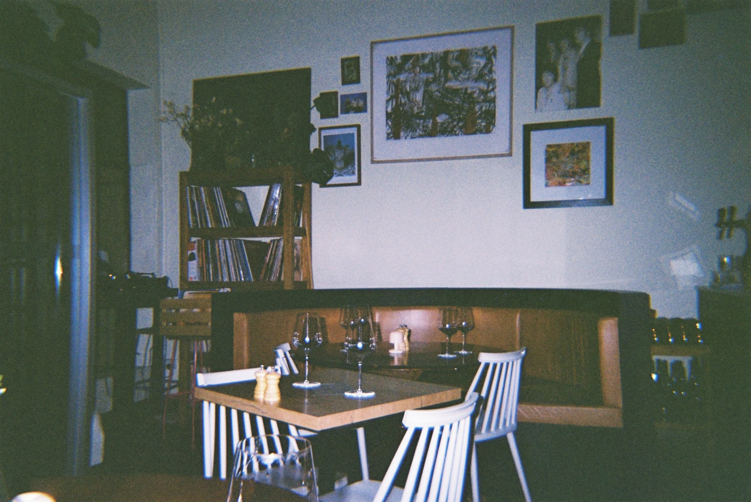A dining room with a wooden table set with wine glasses and a salt and pepper shaker, surrounded by white chairs, with a wall decorated with several framed pictures and a shelf with vinyls and a vase of flowers.