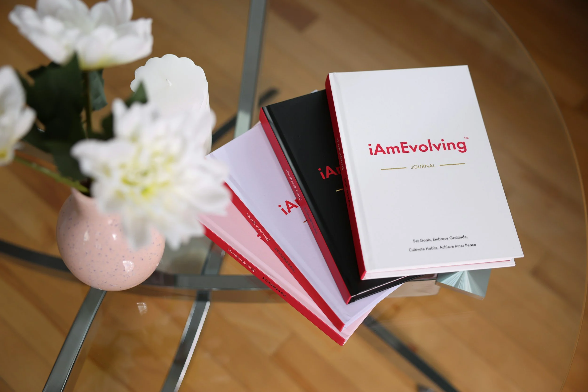 A glass table with a pink vase containing white flowers. Three books titled iAmEvolving Journal stacked on the table.