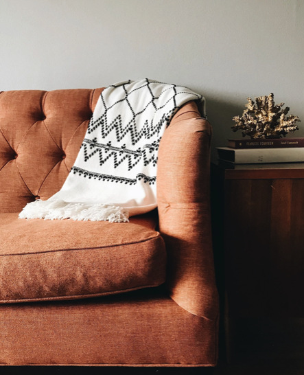 A pinkish-orange tufted sofa with a white and black patterned throw blanket draped over the armrest, next to a dark wooden side table holding a stack of books.